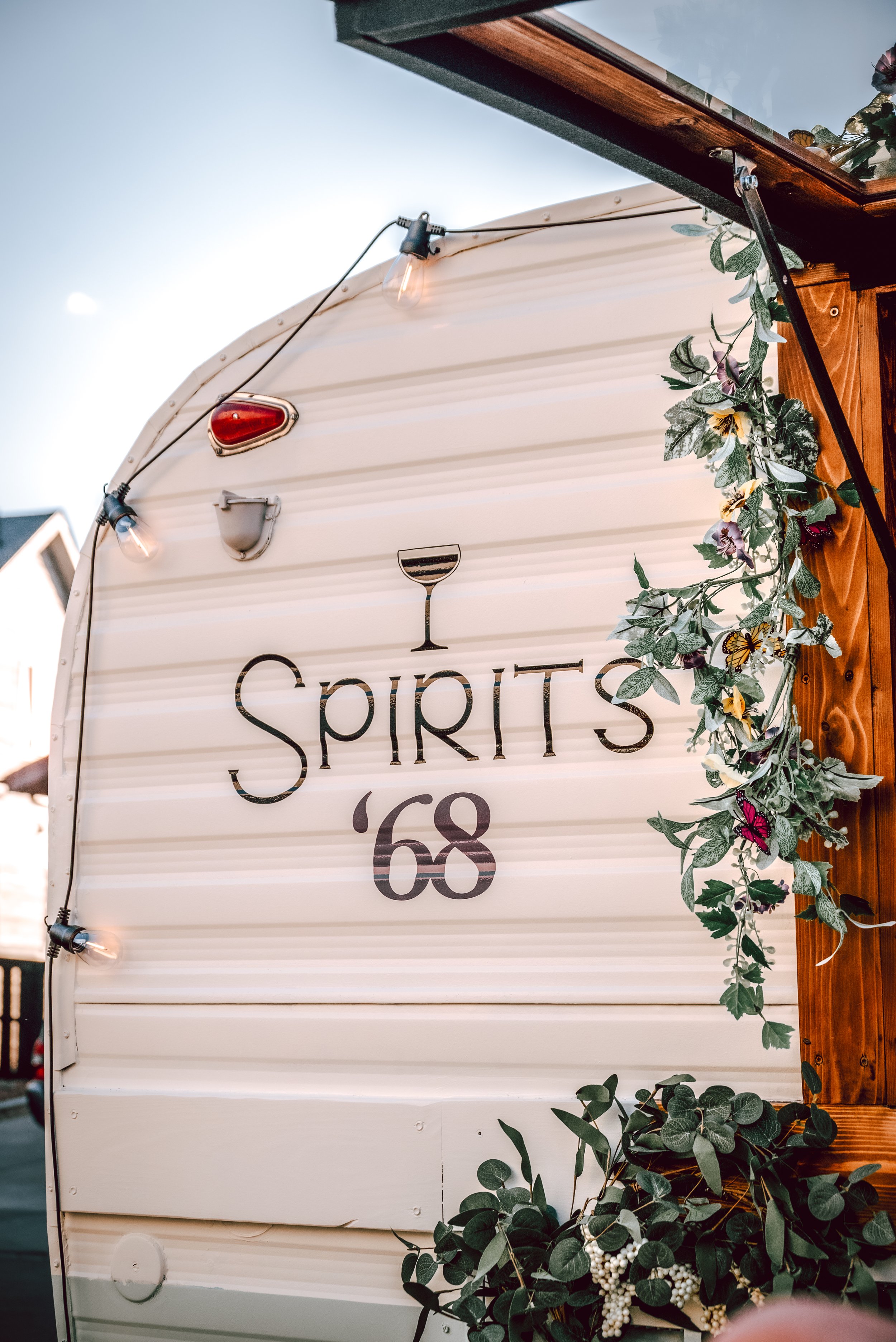 Decorative sign on a vintage camper trailer with the word 'SPIRITS' and the number '68', adorned with string lights and greenery.