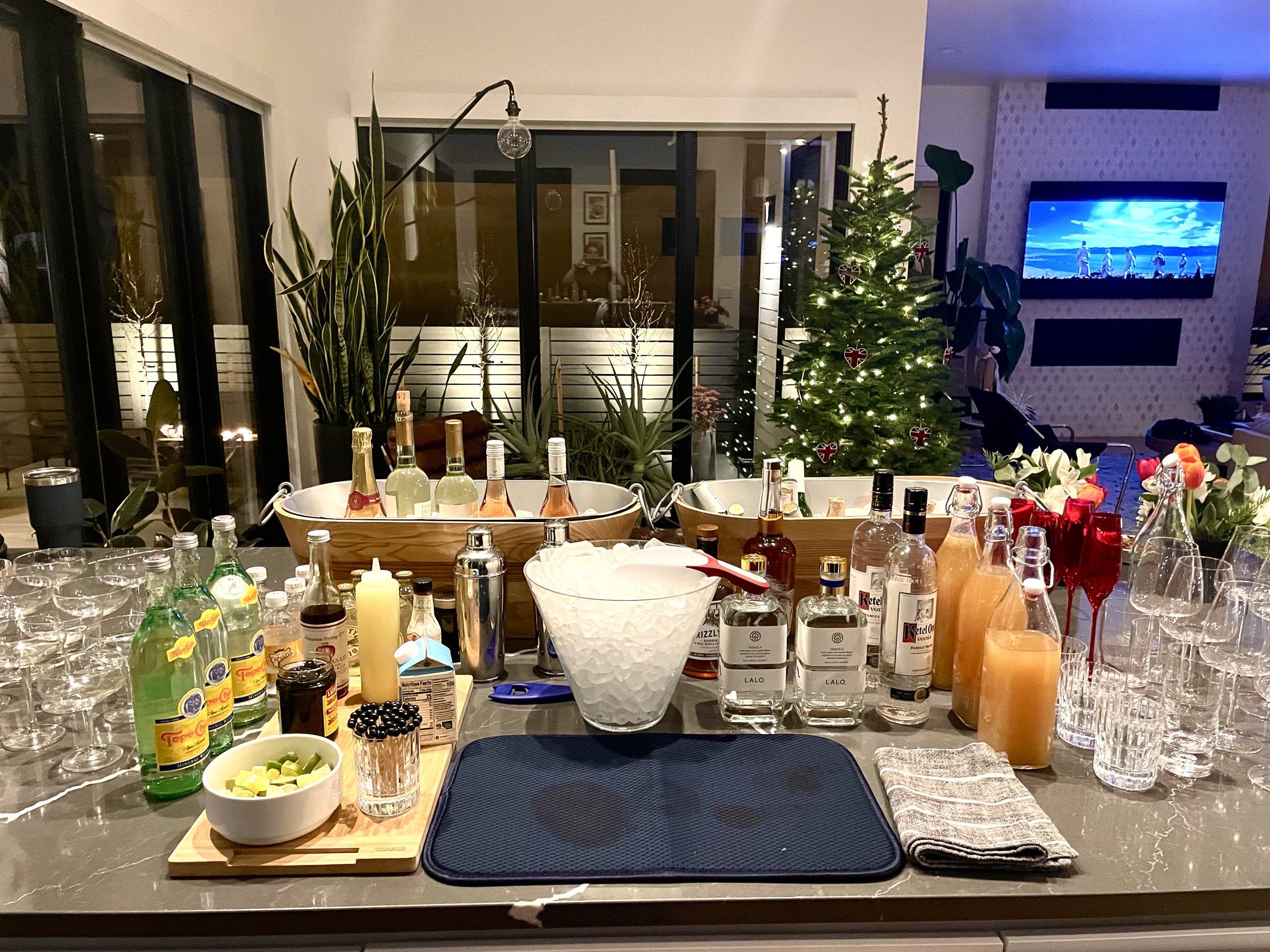 Bar counter with various bottles of alcohol, mixers, glasses, wine, and a bowl of lemon and lime wedges, set for a party with a decorated Christmas tree in the background.