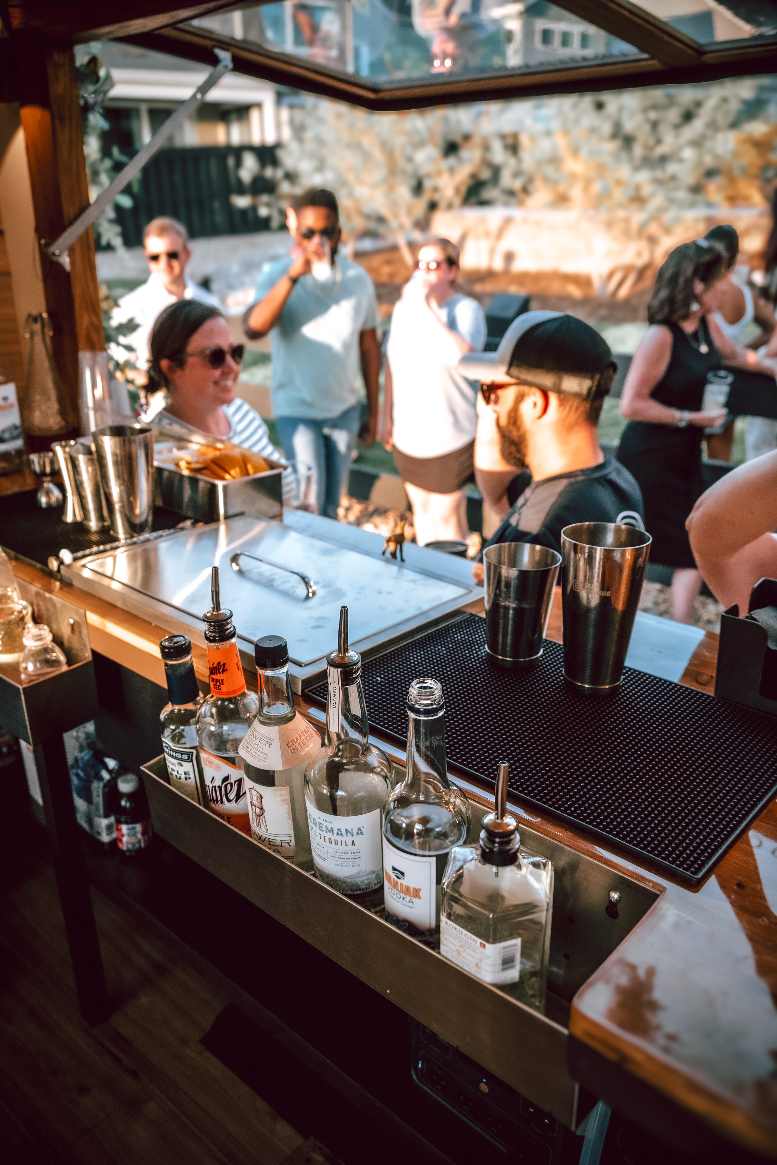 A bar setup with various liquor bottles, bar tools, and metal cups, with people socializing outside in the background during sunset.