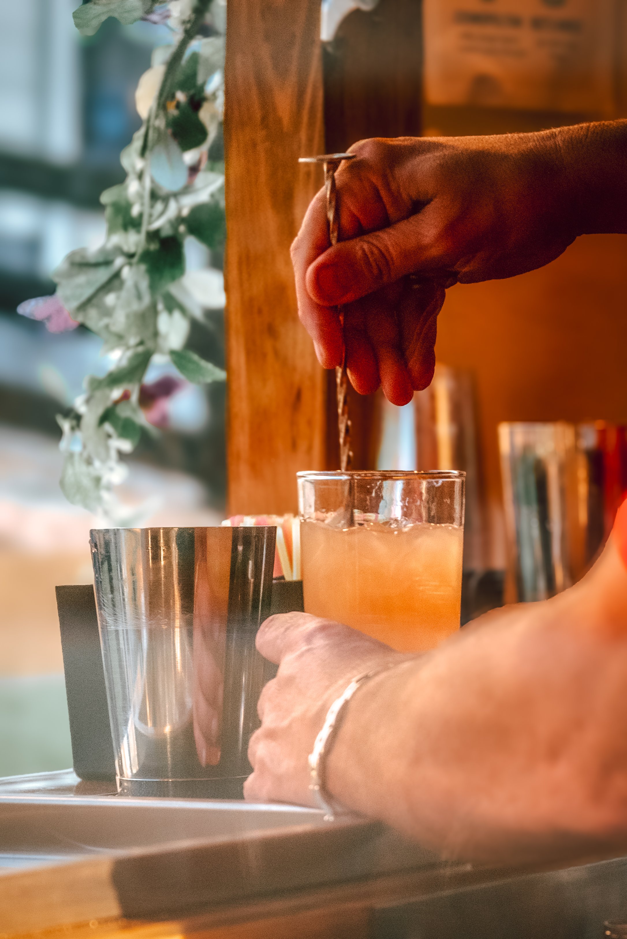 A person is stirring a drink with a metal straw in a glass filled with ice and an orange beverage, while another hand holds a similar drink on a tray.