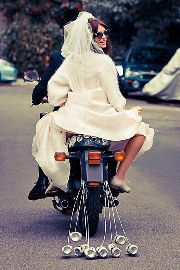 A woman dressed as a bride riding on a motorcycle with a man in the background.