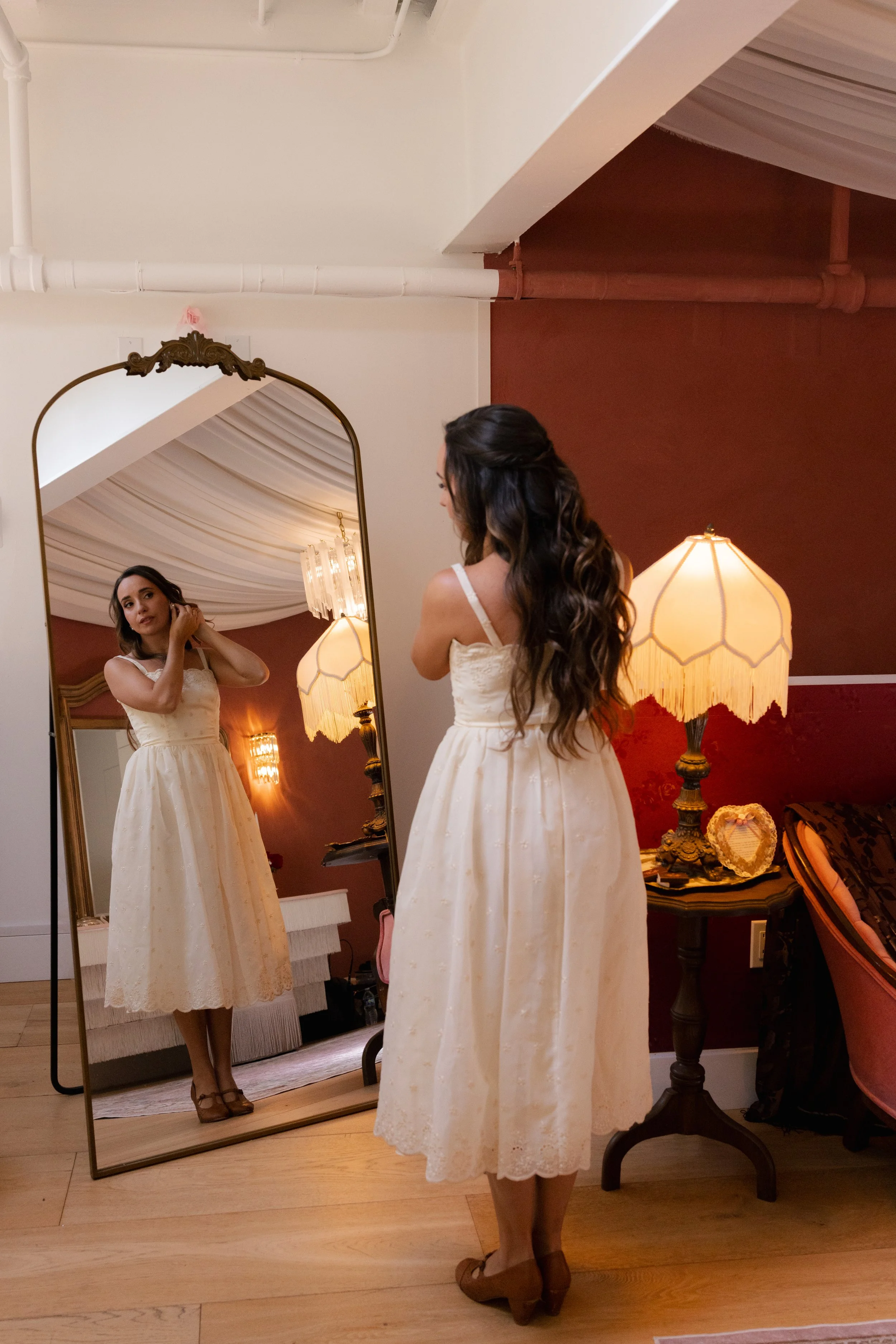 A woman with long, wavy hair wearing a white dress, looking at herself in a tall mirror, adjusting her earrings in a warmly lit room with red walls, a vintage table lamp, and decorative items on a side table.