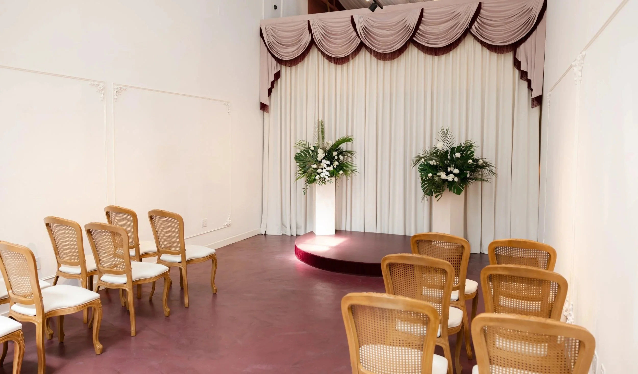An event space setup with rows of wooden chairs with cane backs facing a small stage with a maroon carpet. The stage has large drapes in pink and beige, and there are floral arrangements with white flowers and green leaves on the stage. The room has 