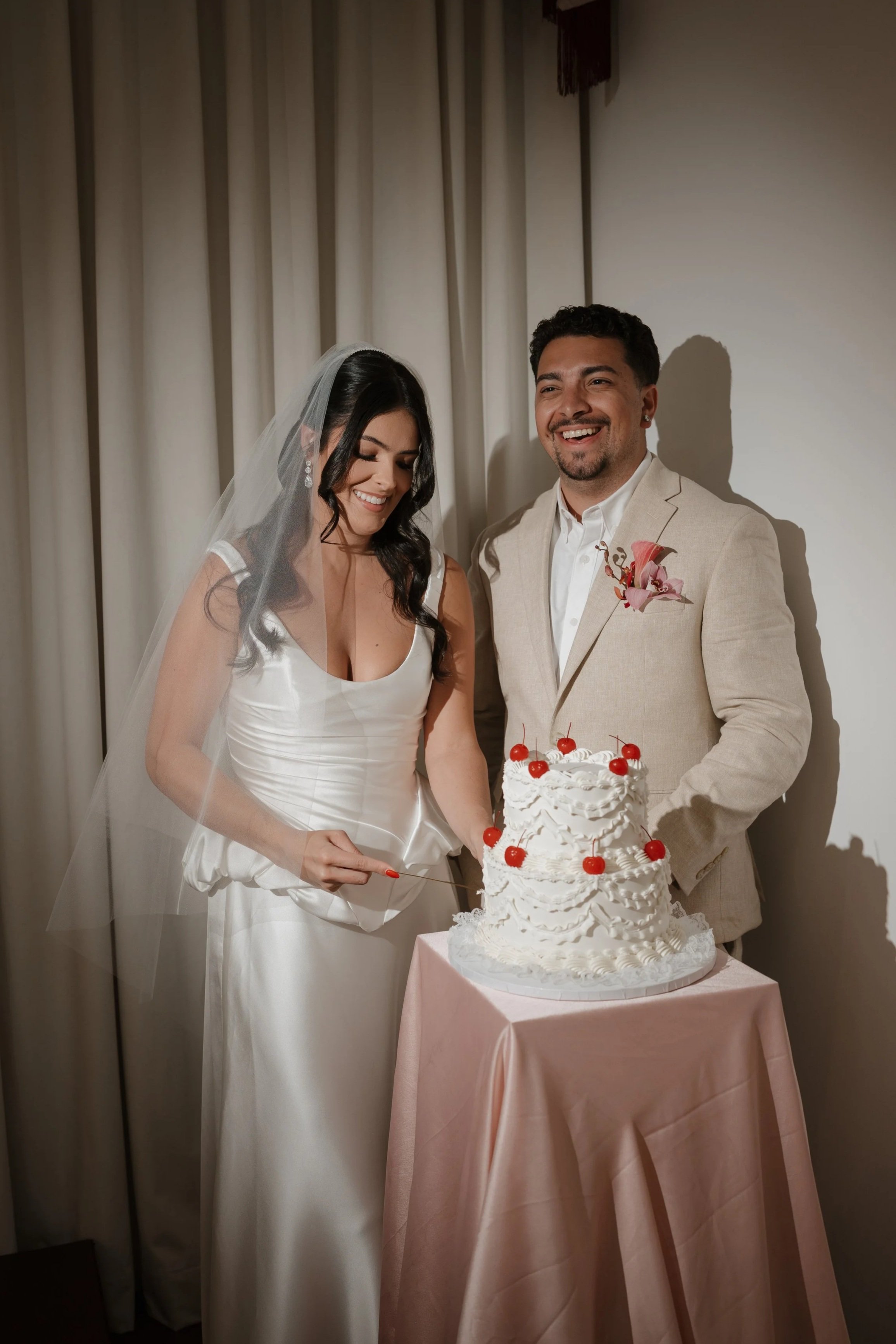 A wedding cake with white frosting and cherries on top, placed on a pink tablecloth. The bride and groom are cutting the cake together.