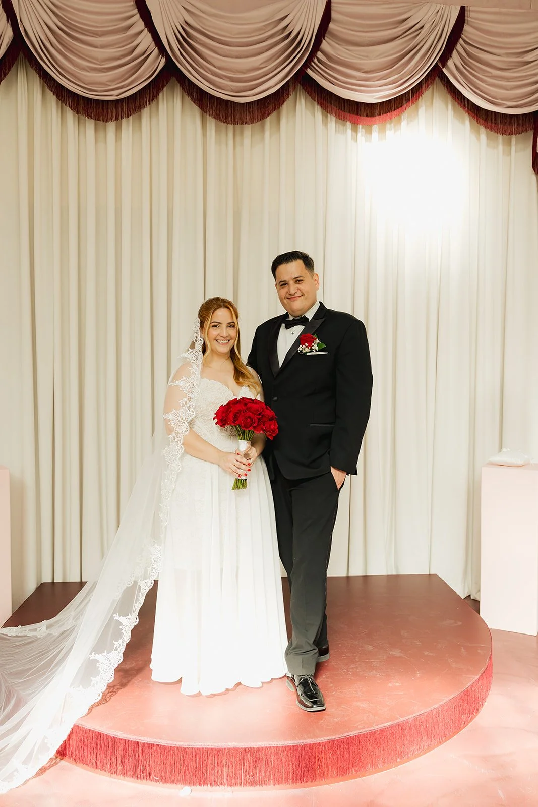 Bridal couple standing on a stage with cream curtains, the bride holding a bouquet of red roses, both dressed in wedding attire, smiling at the camera.