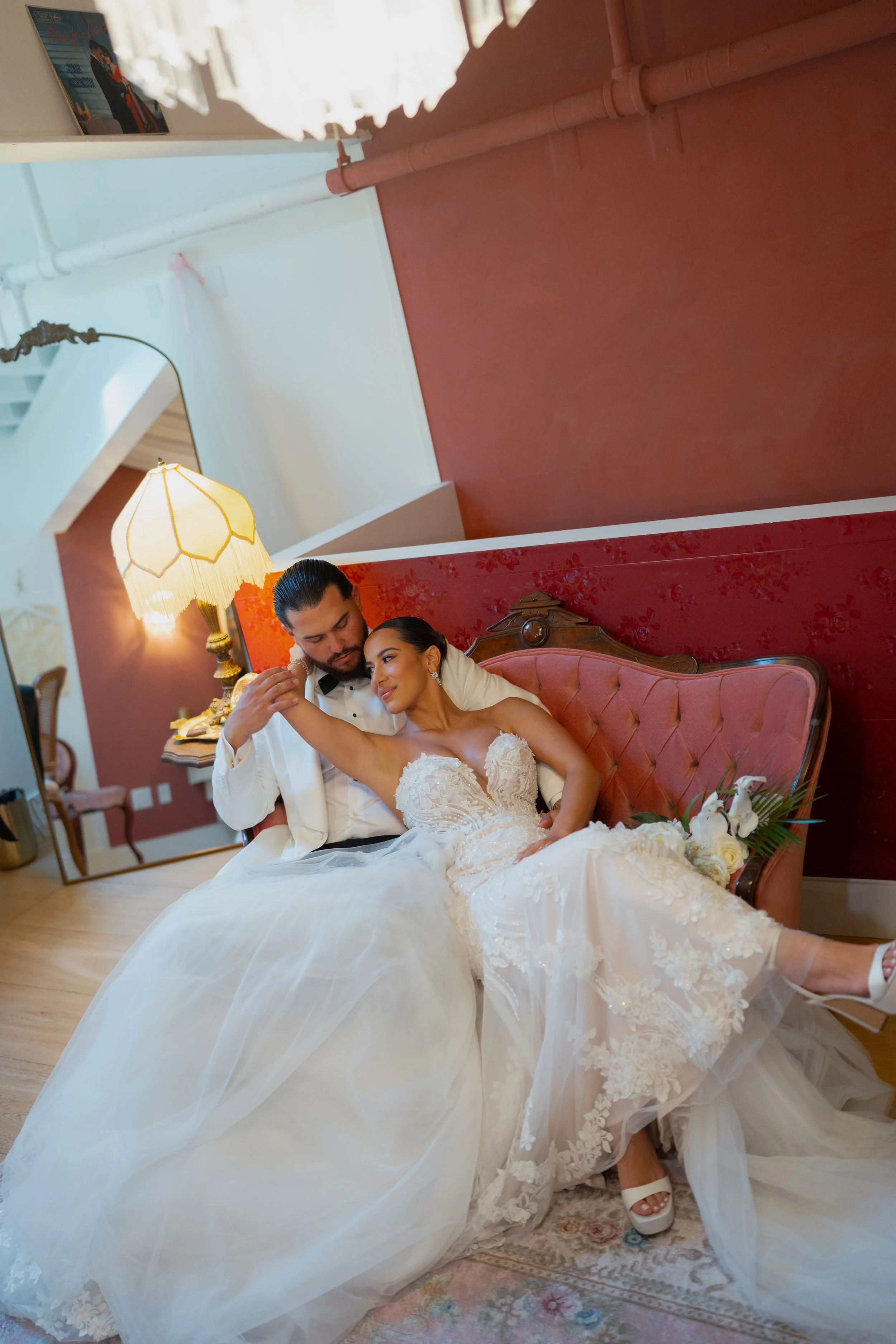 An engaged couple in wedding attire sitting on a vintage pink sofa in a cozy, warmly lit room. The bride is in a white lace wedding dress with a floral bouquet on her lap, and the groom is in a white tuxedo jacket with a black bow tie, gently holding