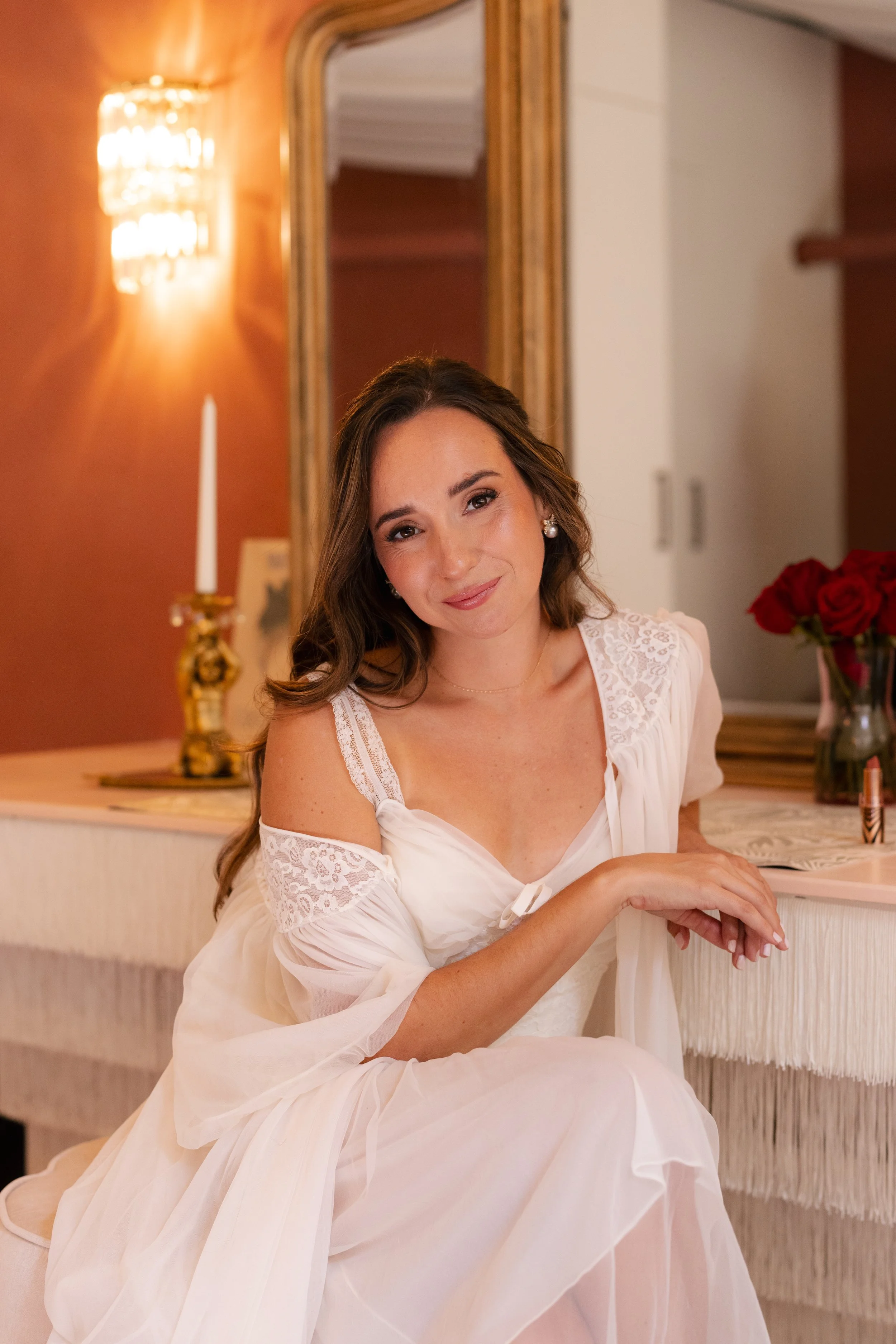 A woman in a white dress sitting at a table in a warmly lit room, with a mirror on the wall behind her and a vase of red roses in the background.