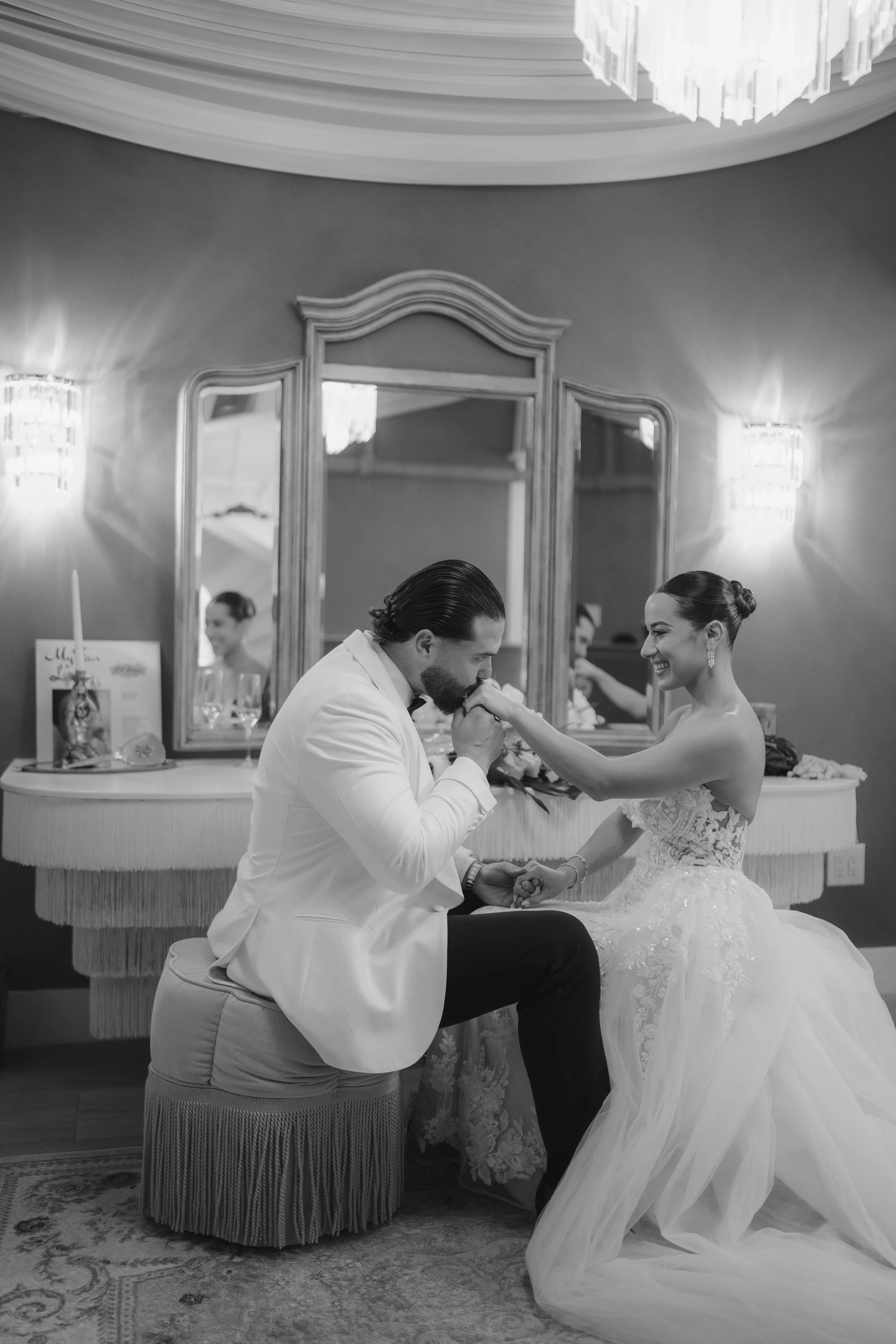 A black and white photo of a wedding ceremony showing a groom kneeling and holding the bride's hand, who is wearing a wedding dress, in front of a mirror. There are reflections of the couple and a woman in the mirror, and the room is decorated with w