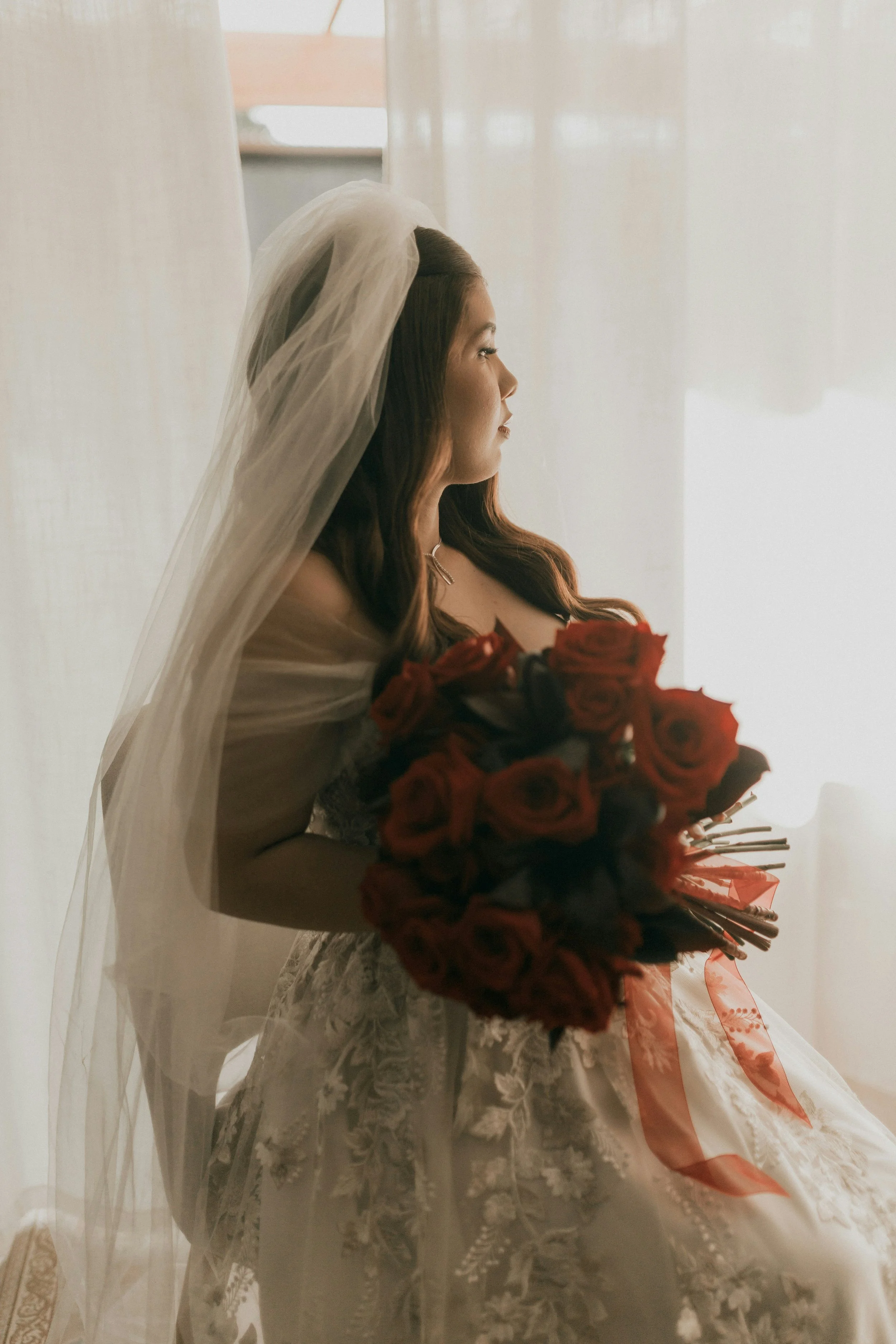 A bride with long brown hair in a white wedding dress and veil, holding a large bouquet of red roses, sitting by a window with sheer white curtains and looking out.