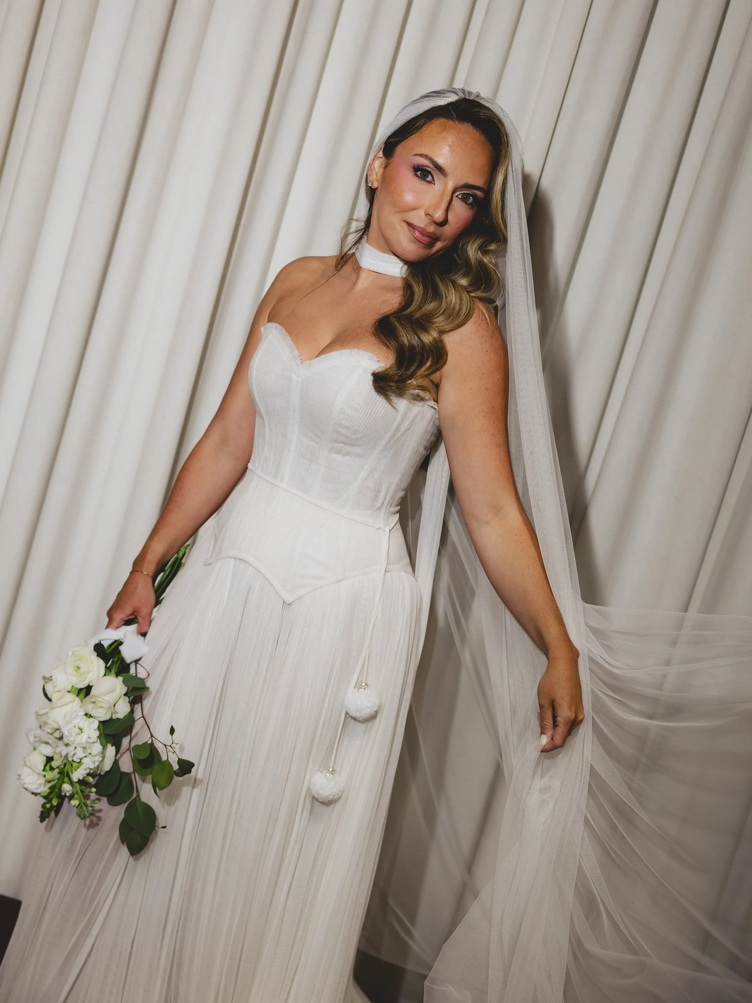 A bride with long blonde hair in a white wedding dress and veil, holding a large bouquet of red roses, sitting by a window with sheer white curtains and looking out.
