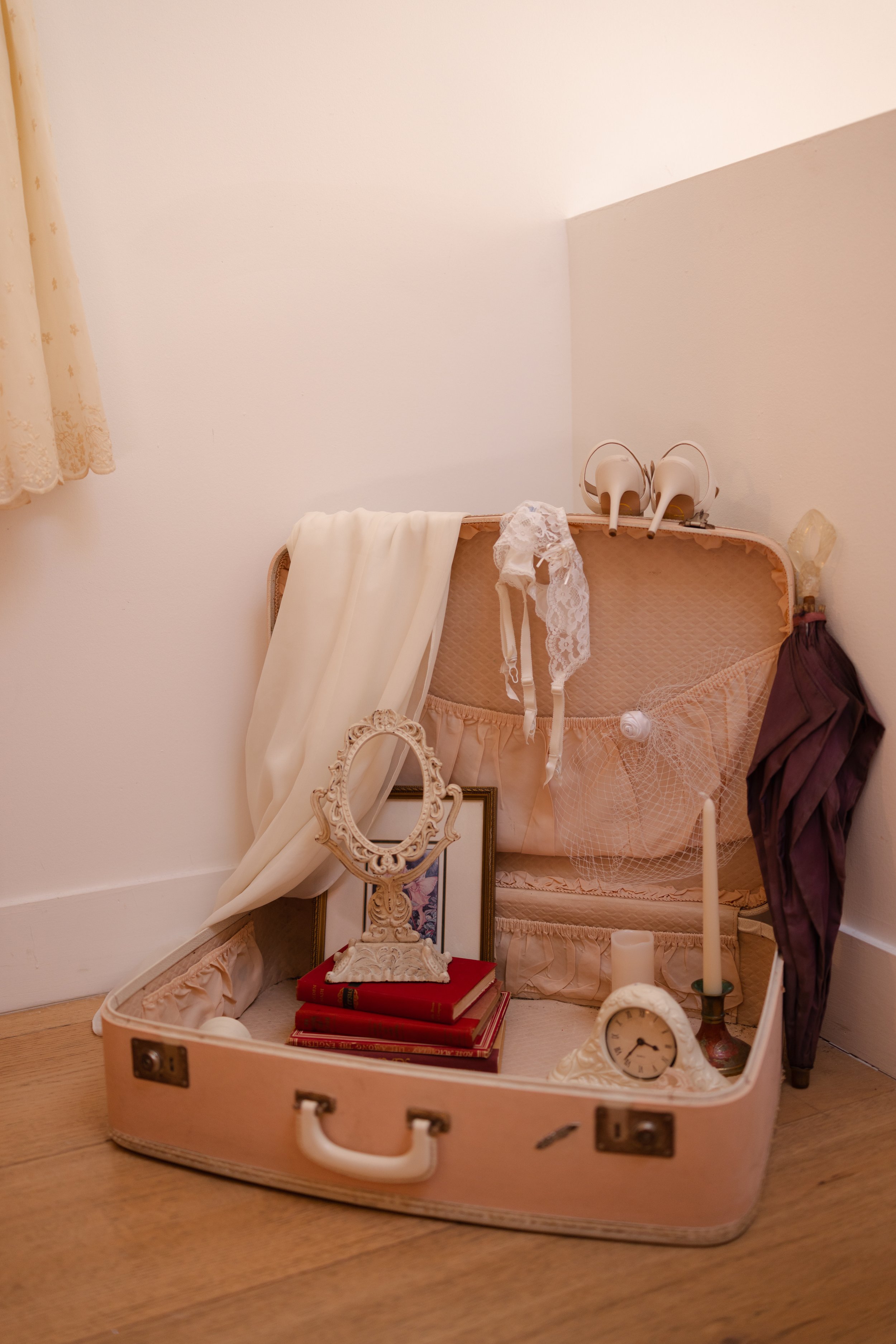 A vintage pink suitcase filled with decorative items such as a white lace garment, a ornate mirror, books, a clock, candles, and various accessories, with a cream-colored curtain and a plain white wall in the background.