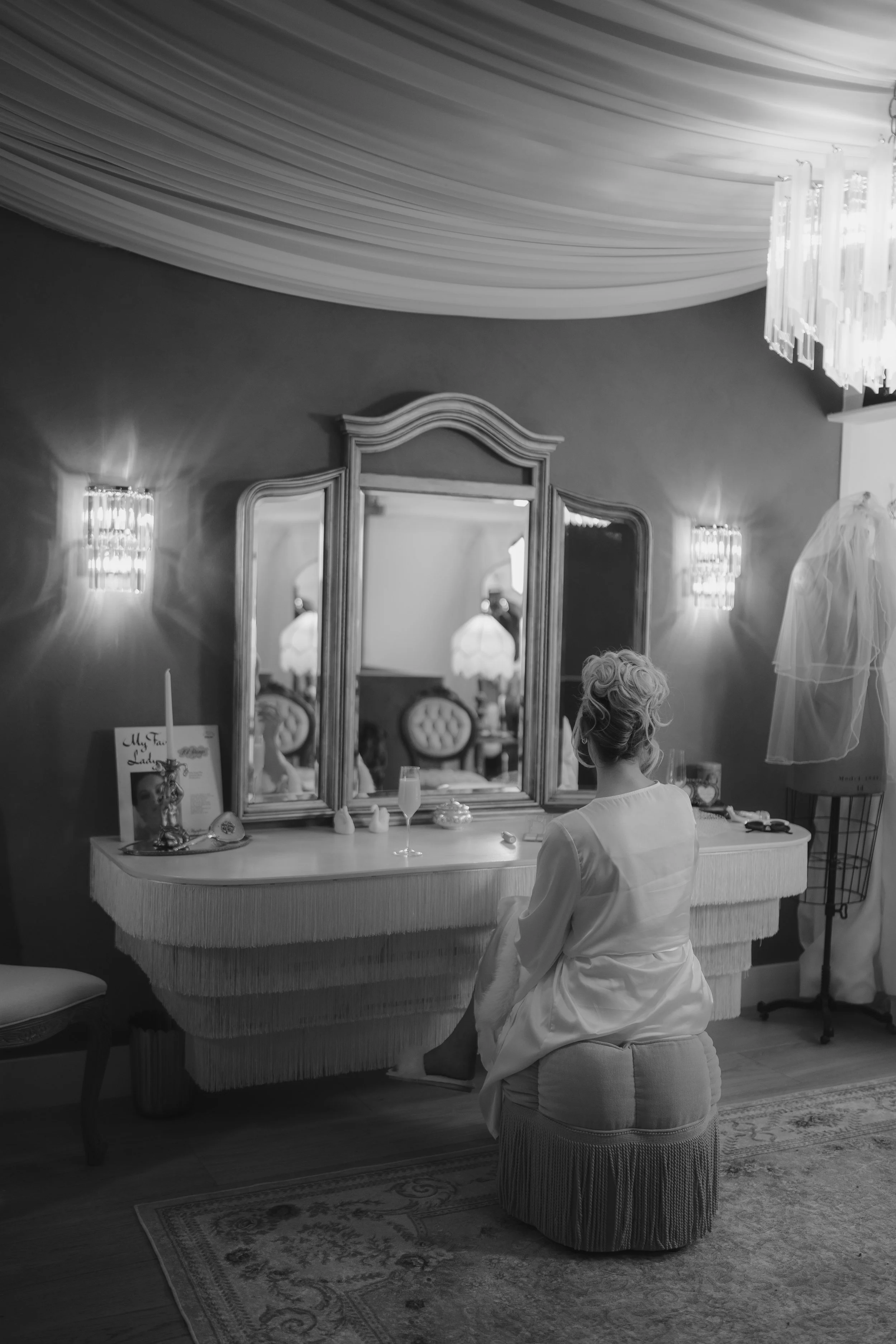 A woman in a white satin robe sitting on a round cushioned stool in front of a dressing table with a large mirror in an elegant room decorated with chandeliers, wall sconces, and a draped ceiling. The room has a vintage style with ornate furniture an