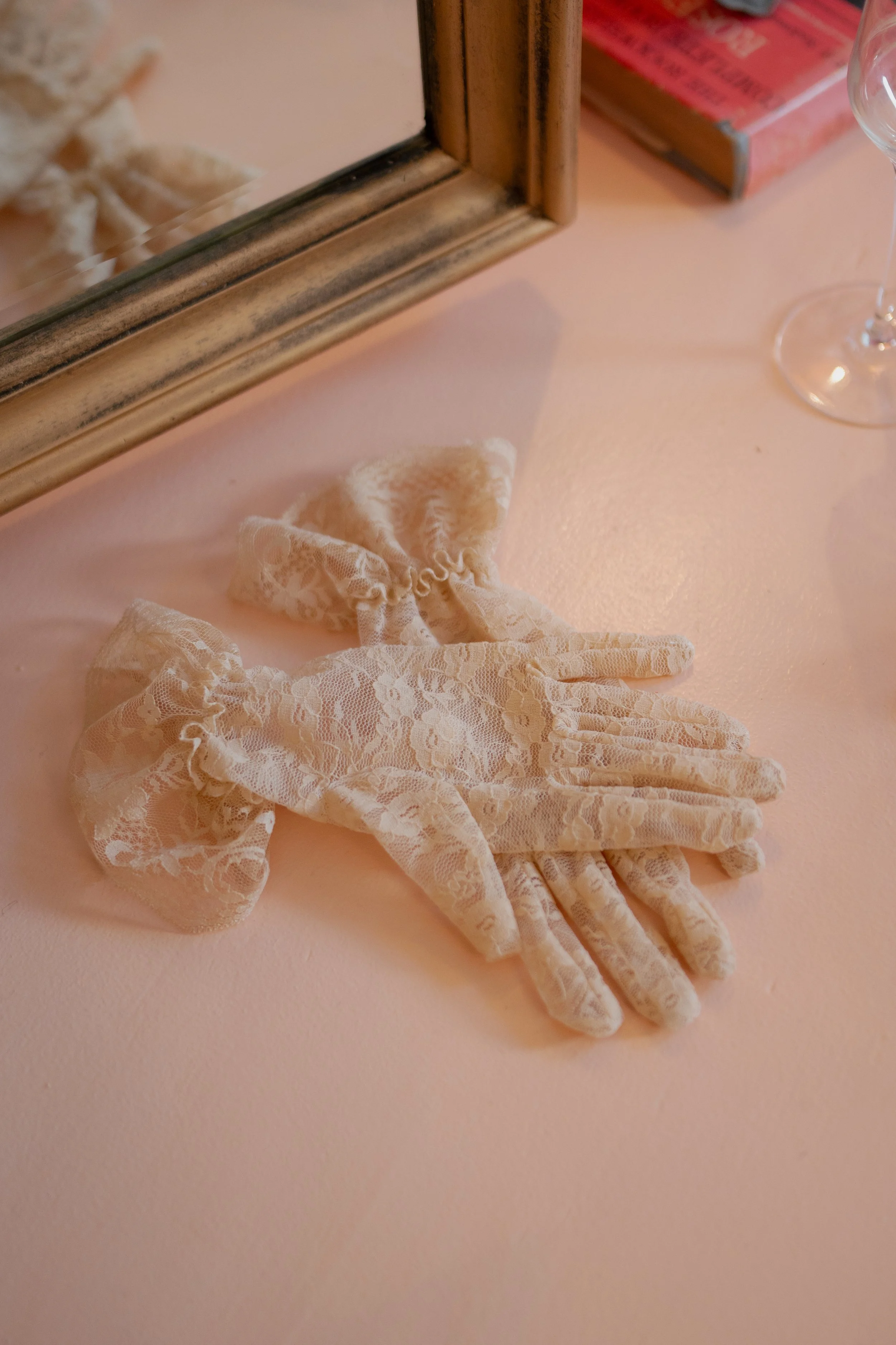 A pair of white lace gloves placed on a pink surface with a mirror, a glass, and a box in the background.
