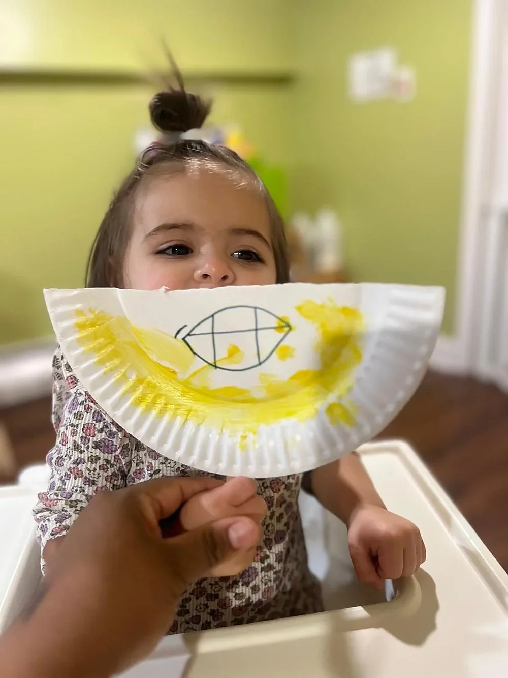 Young girl with brown hair tied in a bun, sitting in a high chair, holding a paper plate with yellow paint and a drawing, as a person offers it to her, in a room with green walls.