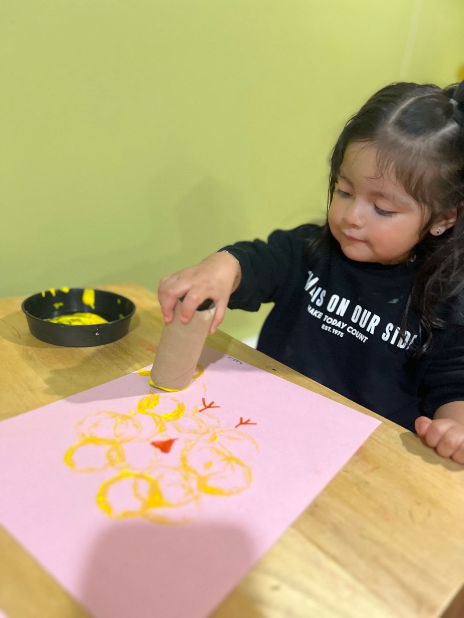 A young girl creating a craft project on pink paper, using a cardboard tube and yellow paint, with a black paint dish nearby