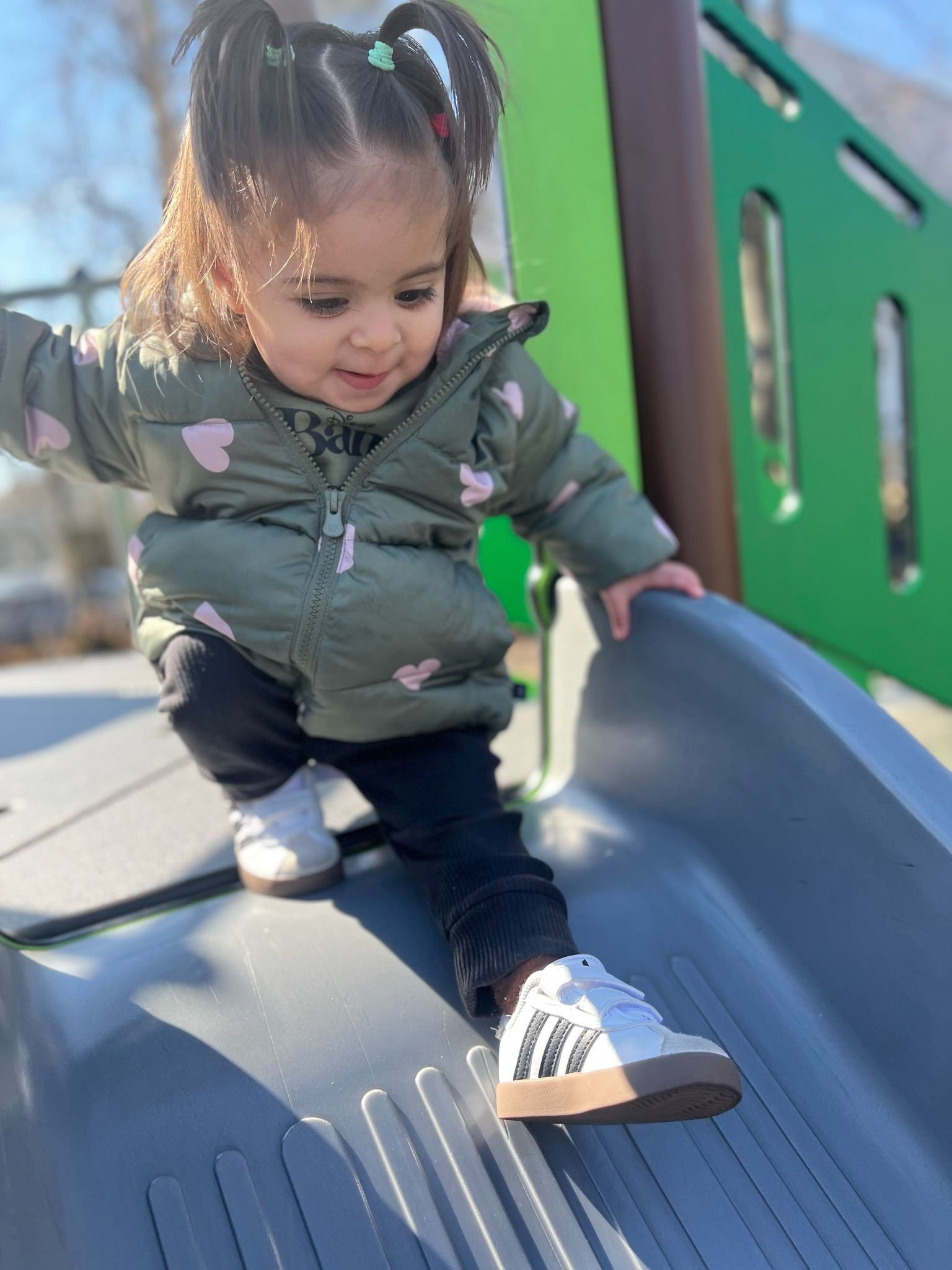 A young girl with pigtails wearing a grey jacket with pink hearts, black pants, and white sneakers, playing on a slide at a playground.