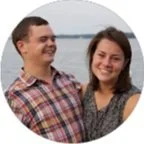 A smiling man and woman standing outdoors near a body of water, both wearing casual clothing.