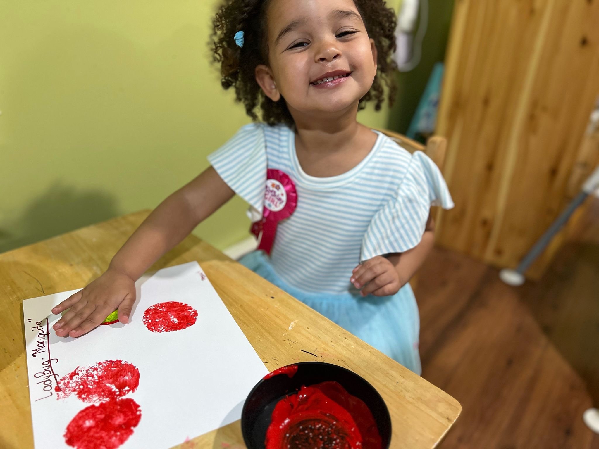 A young girl with curly hair and a big smile displays red handprints on a piece of paper, proud of her artwork, sitting at a wooden table.