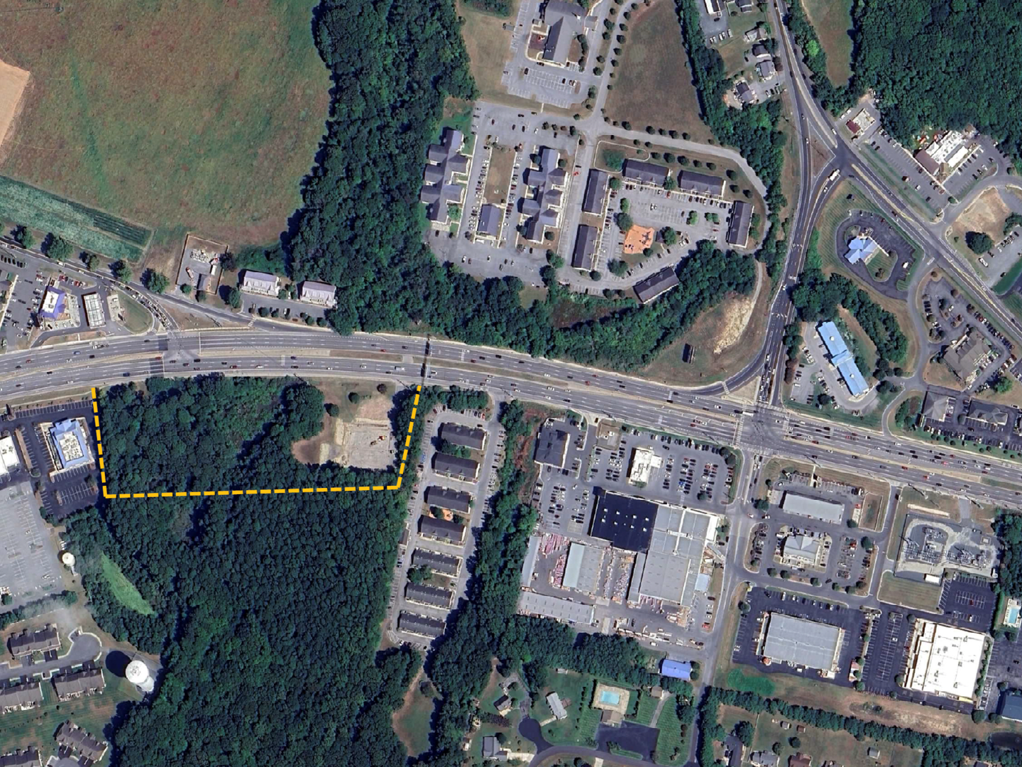 Aerial view of an urban area showing roads, parking lots, buildings, and a green wooded area outlined with dashed yellow lines.