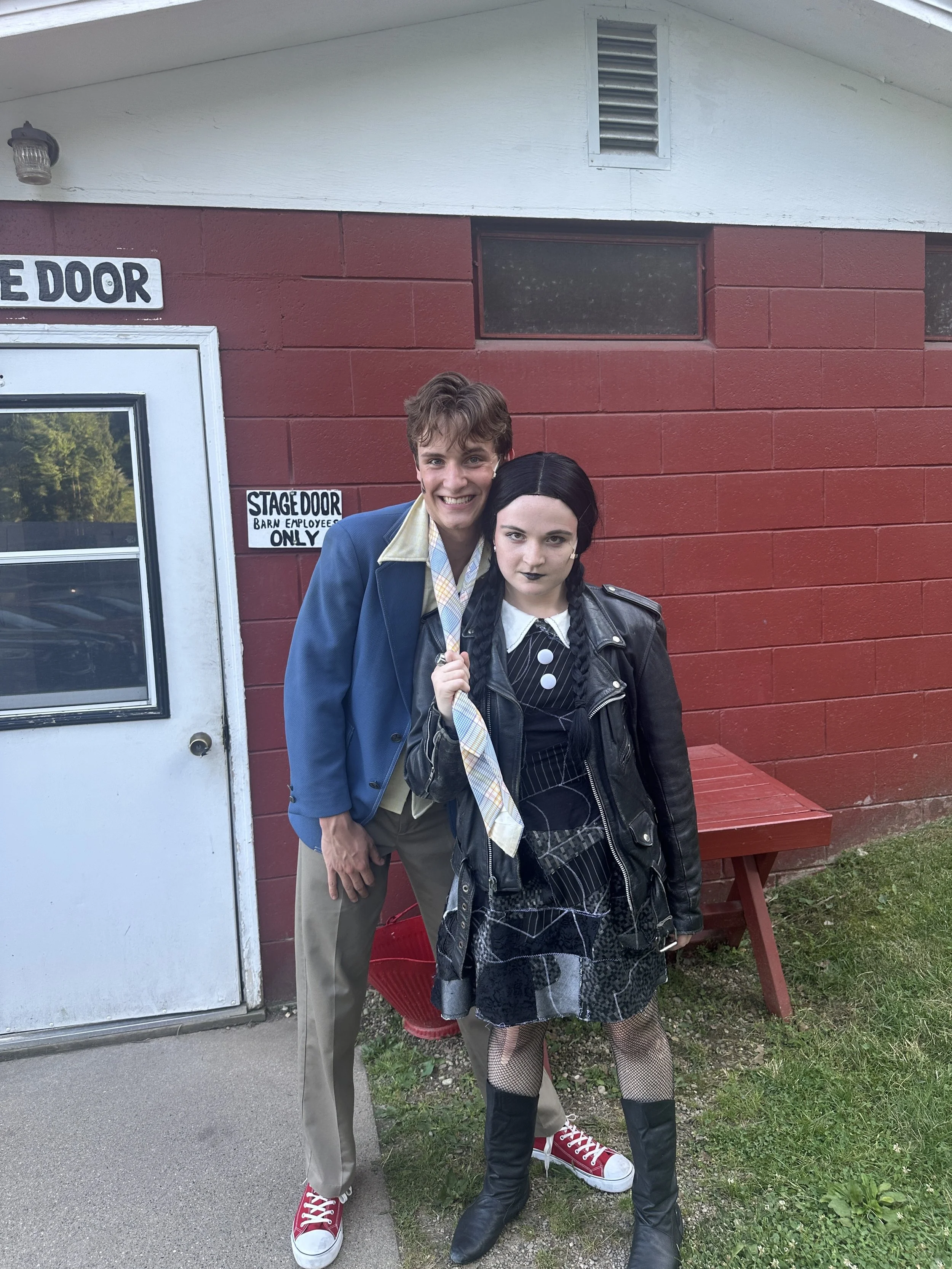 Two people standing outside near a red brick building, one man in a blue blazer and beige pants, the other woman dressed in dark clothing with fishnet stockings and fishnet gloves, both smiling and posing for the photo.