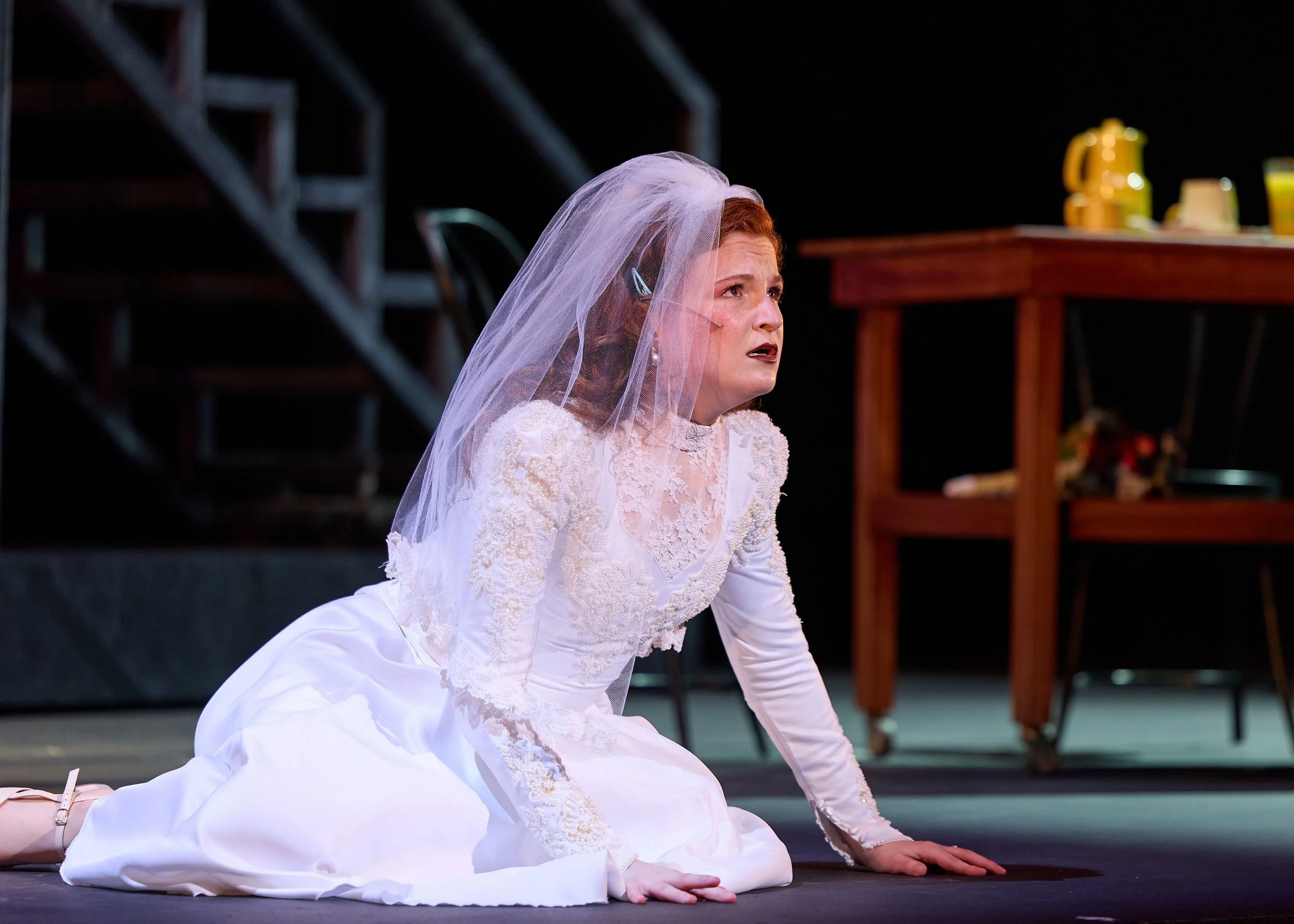 A woman in a wedding dress and veil kneels on stage with her hands on the ground, looking up with a distressed expression.
