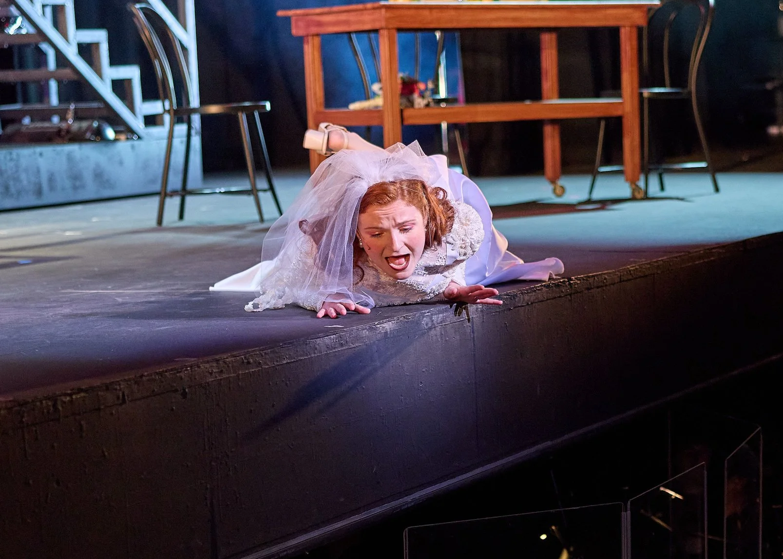 A woman dressed in a wedding gown and veil is lying on the stage floor, appearing distressed, with her face and hands pressed against the stage surface.