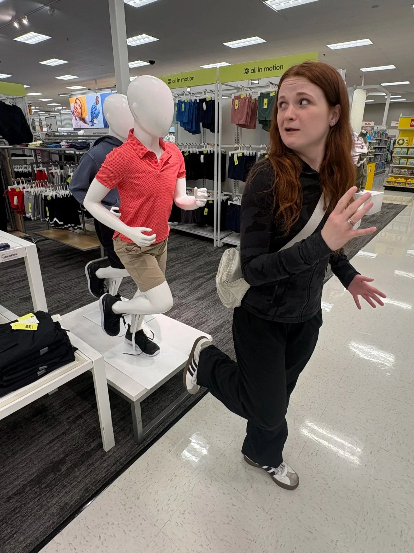 A woman with red hair and black clothing making a surprised or confused expression in a retail store, standing next to a mannequin dressed in red, who appears to be running or in a dynamic pose.