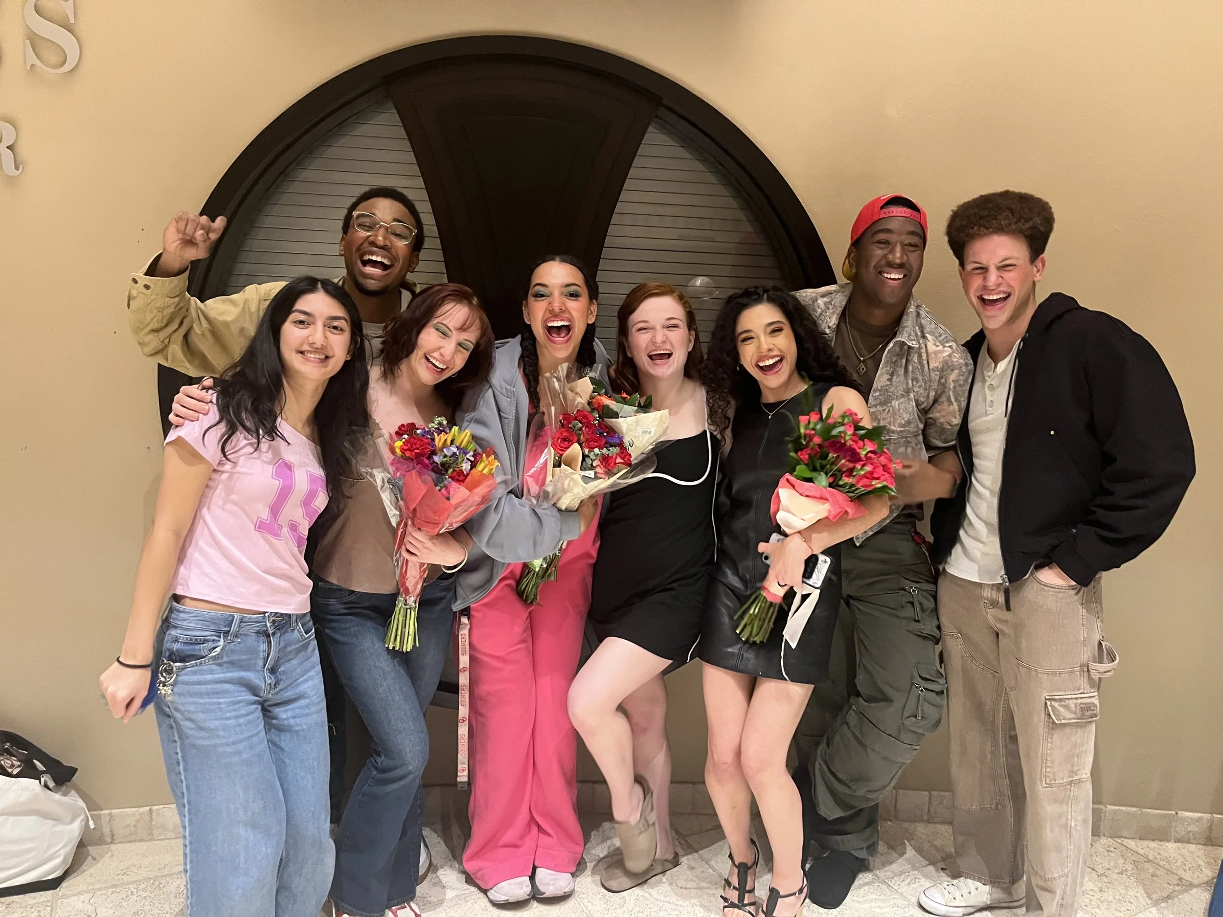 Group of diverse young people celebrating, smiling, holding bouquets of flowers, standing in front of a window with a decorative wall behind them, some wearing casual clothes, one person in a red baseball cap.