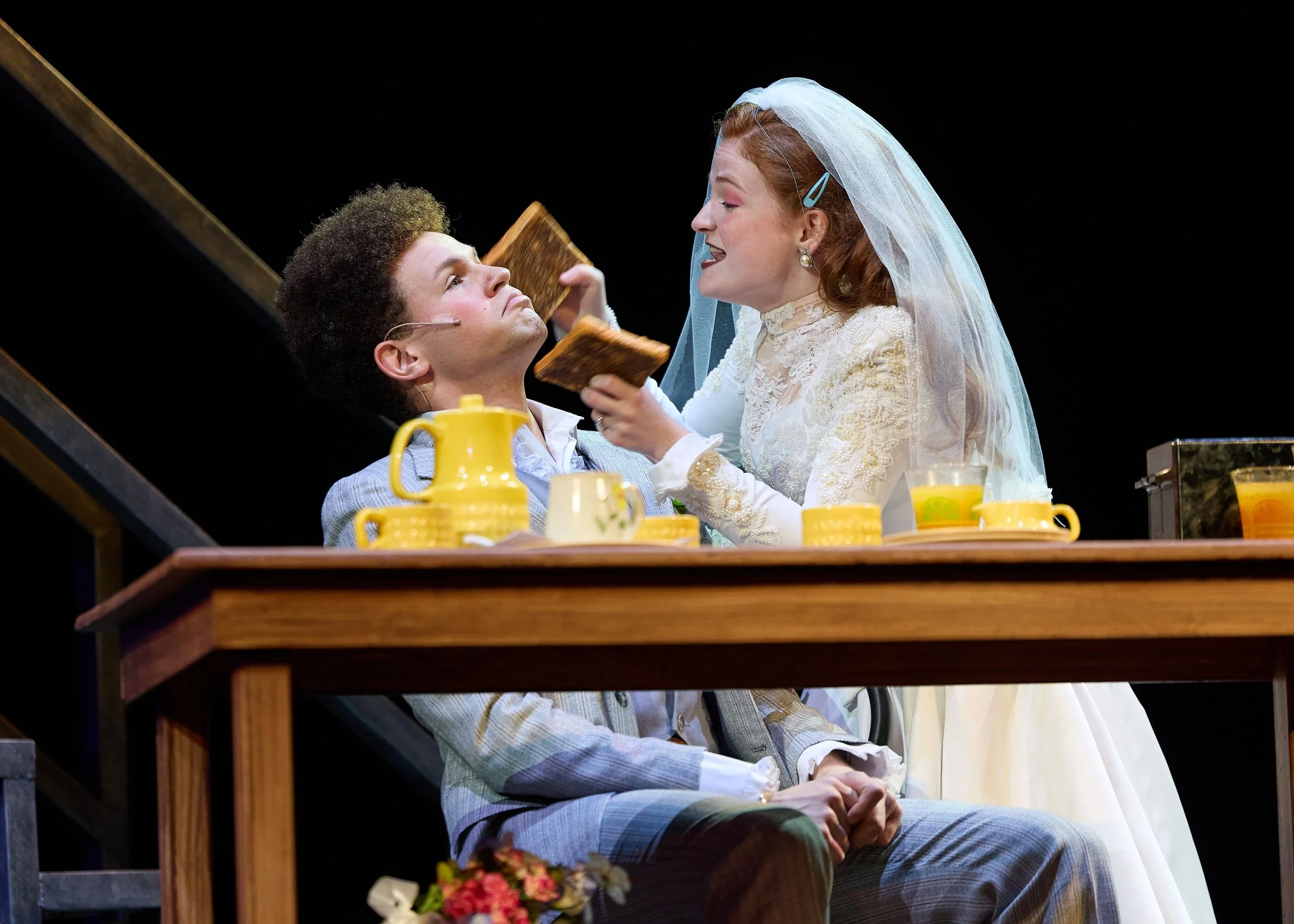 A theatrical scene with a man sitting at a table and a woman in a wedding dress leaning toward him, holding a piece of toast, with yellow teacups and a yellow pitcher on the table, set against a black background.