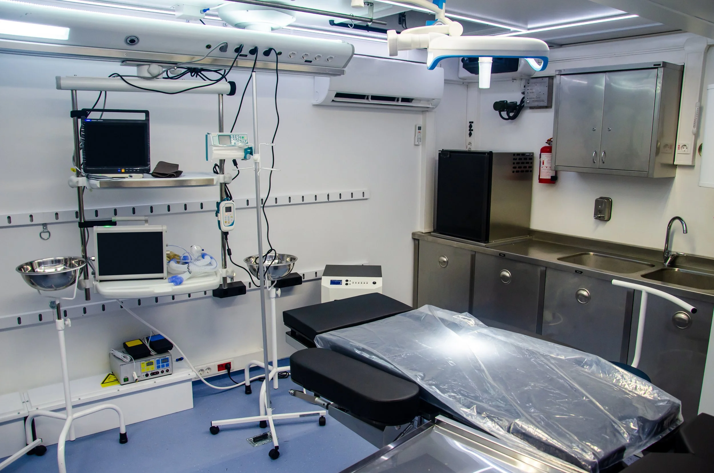 Hospital operating room with medical equipment, a surgical table covered in clear plastic, and various monitors and supplies organized on shelves.