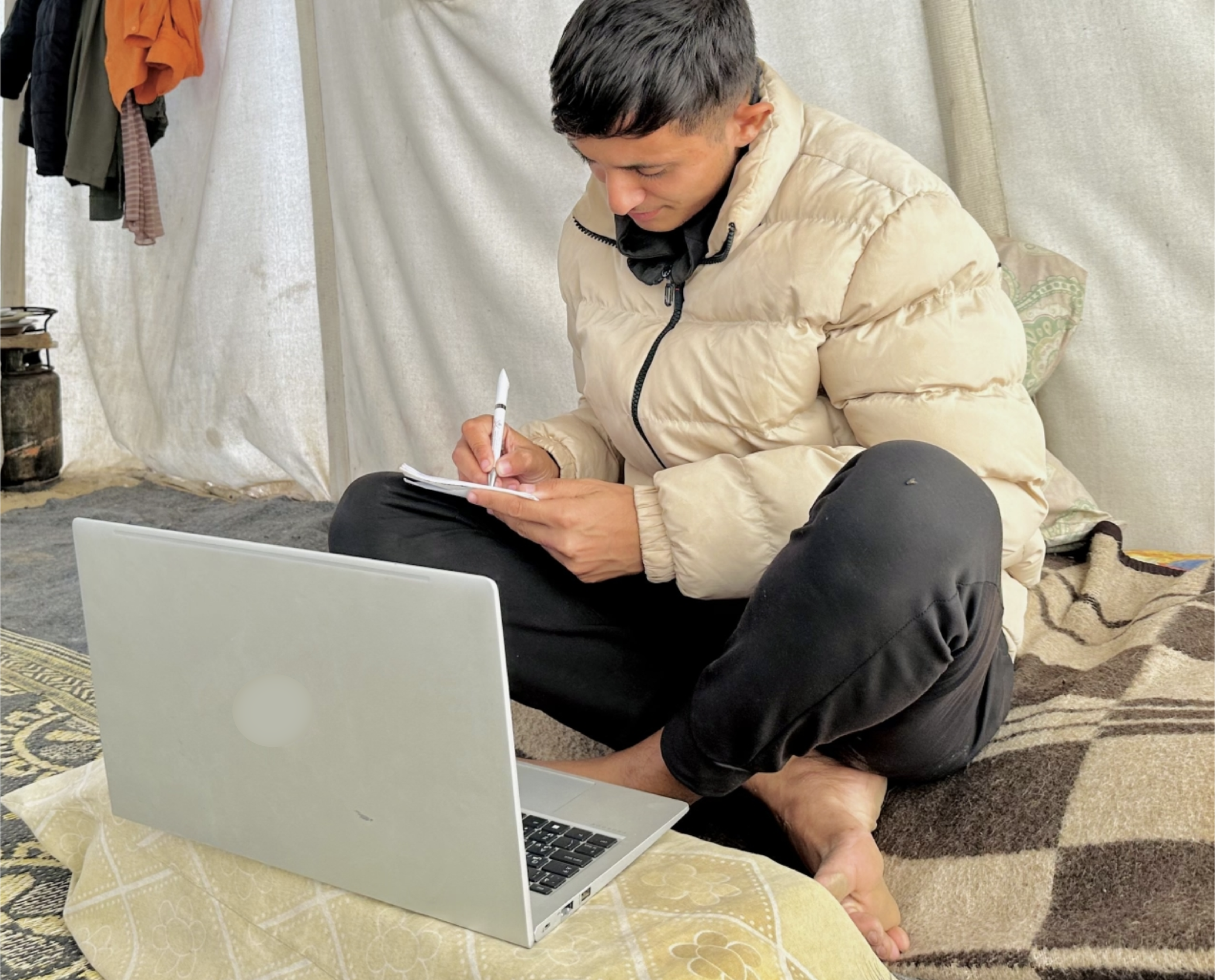 A young man sitting on the floor with a laptop in front of him, writing on a notepad with a pen, inside a makeshift tent or shelter, wearing a beige puffer jacket and black pants.
