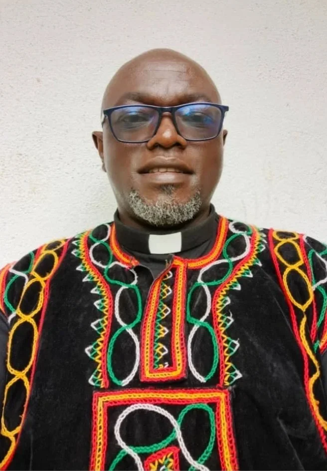 A man wearing glasses and a black clerical collar with a white tab, dressed in a traditional African embroidered shirt with red, yellow, green, and white patterns, standing against a plain light-colored wall.