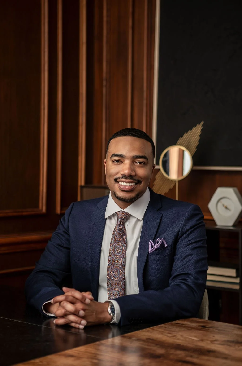 A man in a dark blue suit and colorful tie sitting at a wooden desk, smiling in a professional office with dark wood paneling and modern decor.