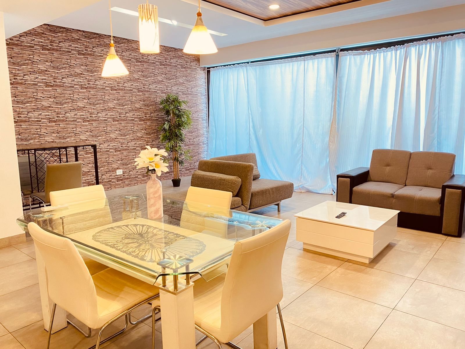 Living room with beige tiled floor, glass dining table with cream-colored chairs, brown sofas, white coffee table, blue curtains, and a textured stone accent wall with hanging pendant lights.