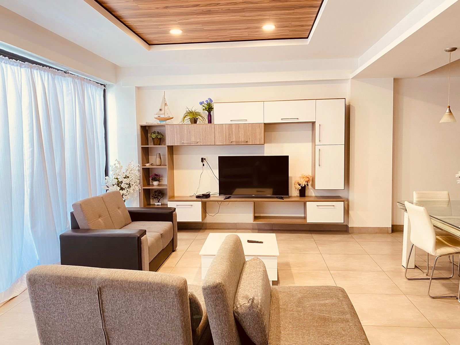 Modern living room with beige and black sofa, white coffee table, and a wall-mounted TV with wooden and white storage units, near a large window with white curtains.