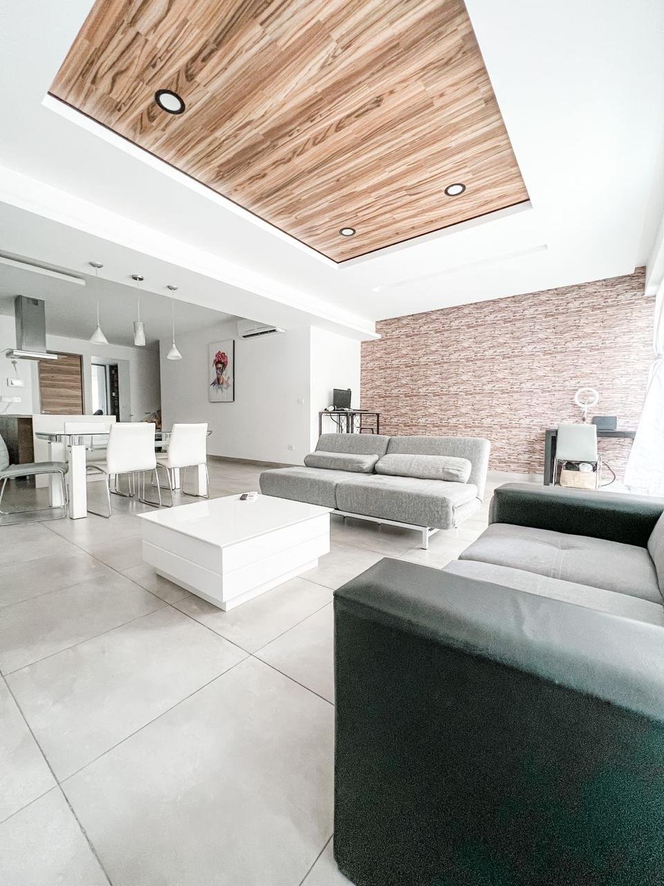 Modern living room with gray and black sofas, white coffee table, tiled floor, partial view of dining area with white chairs, brick accent wall, wooden ceiling panel, and minimal decor.