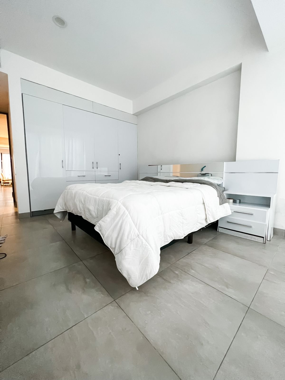 A minimalistic white bedroom with a bed, built-in white wardrobe, and tiled floor.