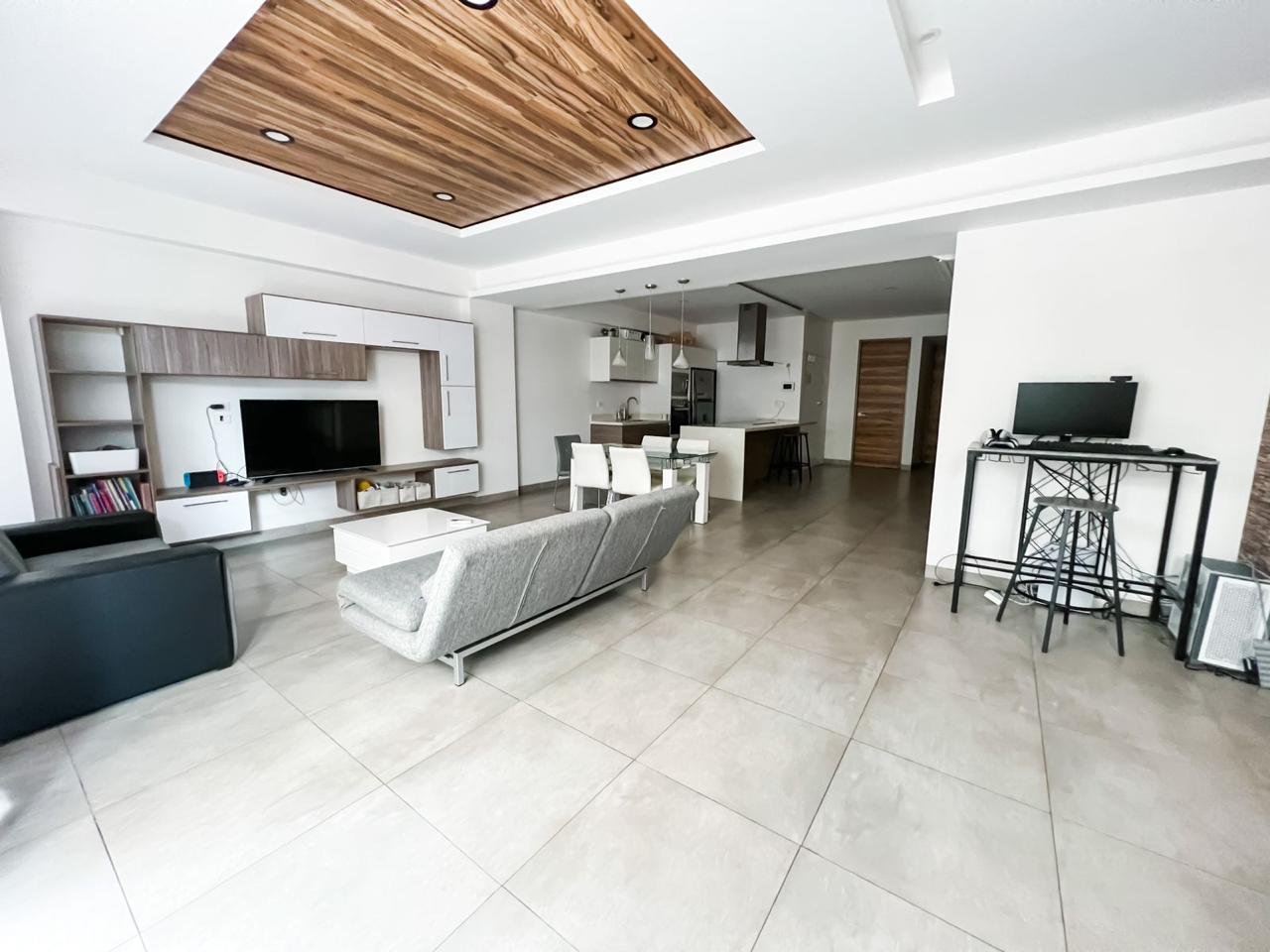 Open-concept living room and kitchen with modern decor, beige tile flooring, white walls, a ceiling with recessed lighting and wood accents, a flat-screen TV, a gray sofa, white dining table with chairs, and small black desk with a computer.