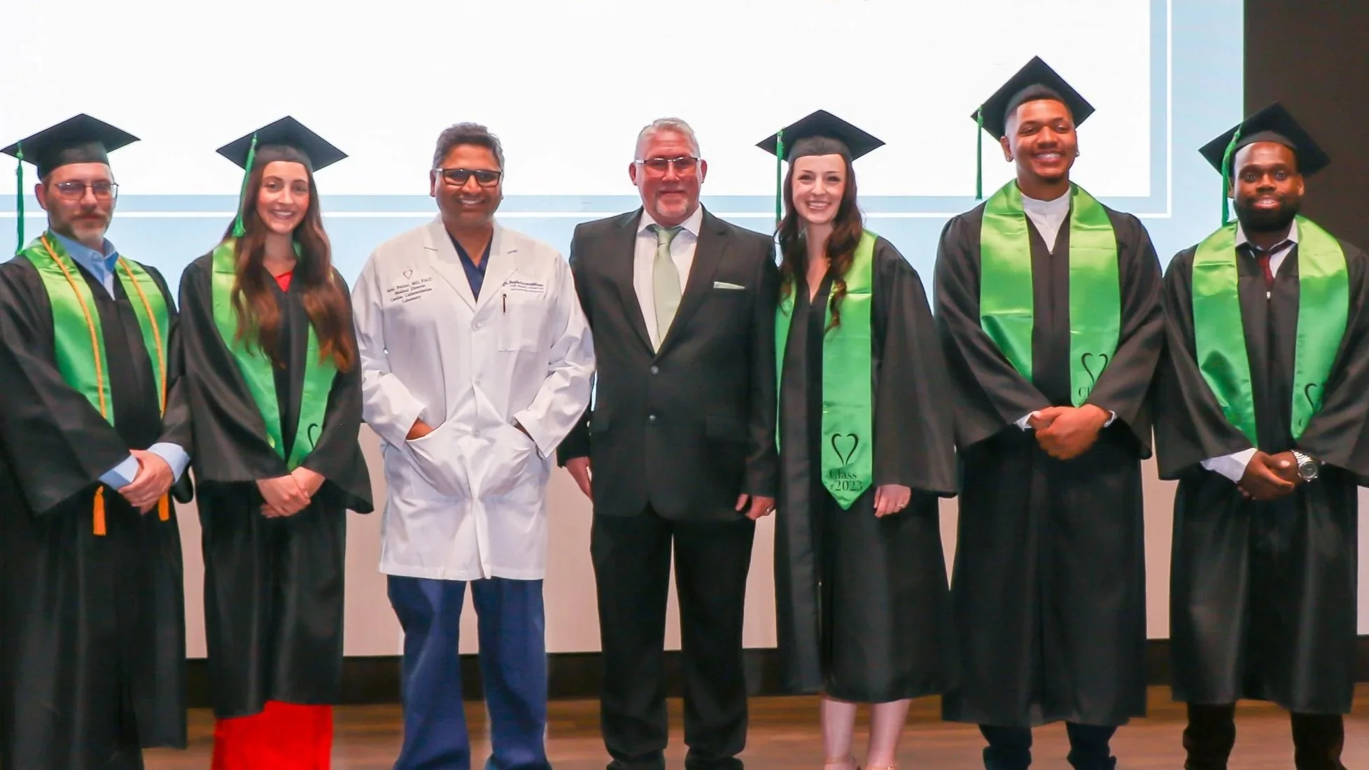 Group of 5 Cardiovascular Institute - Invasive Cardiovascular Technology Perfusion program graduates in caps and gowns standing indoors in front of Baylor Scott & White The Heart Hospital - Plano background image.