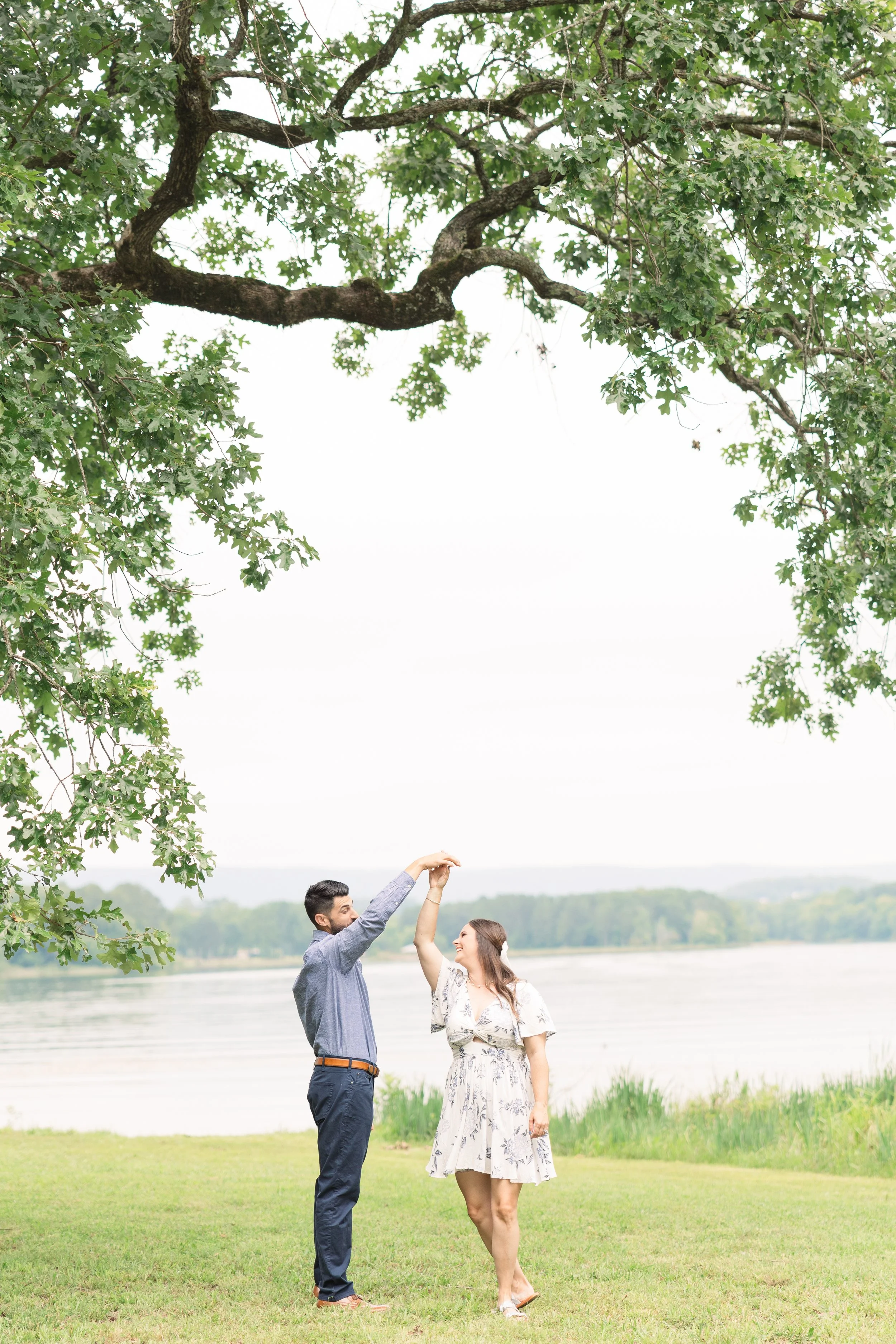 A couple dancing outdoors near a river, framed by large leafy trees.