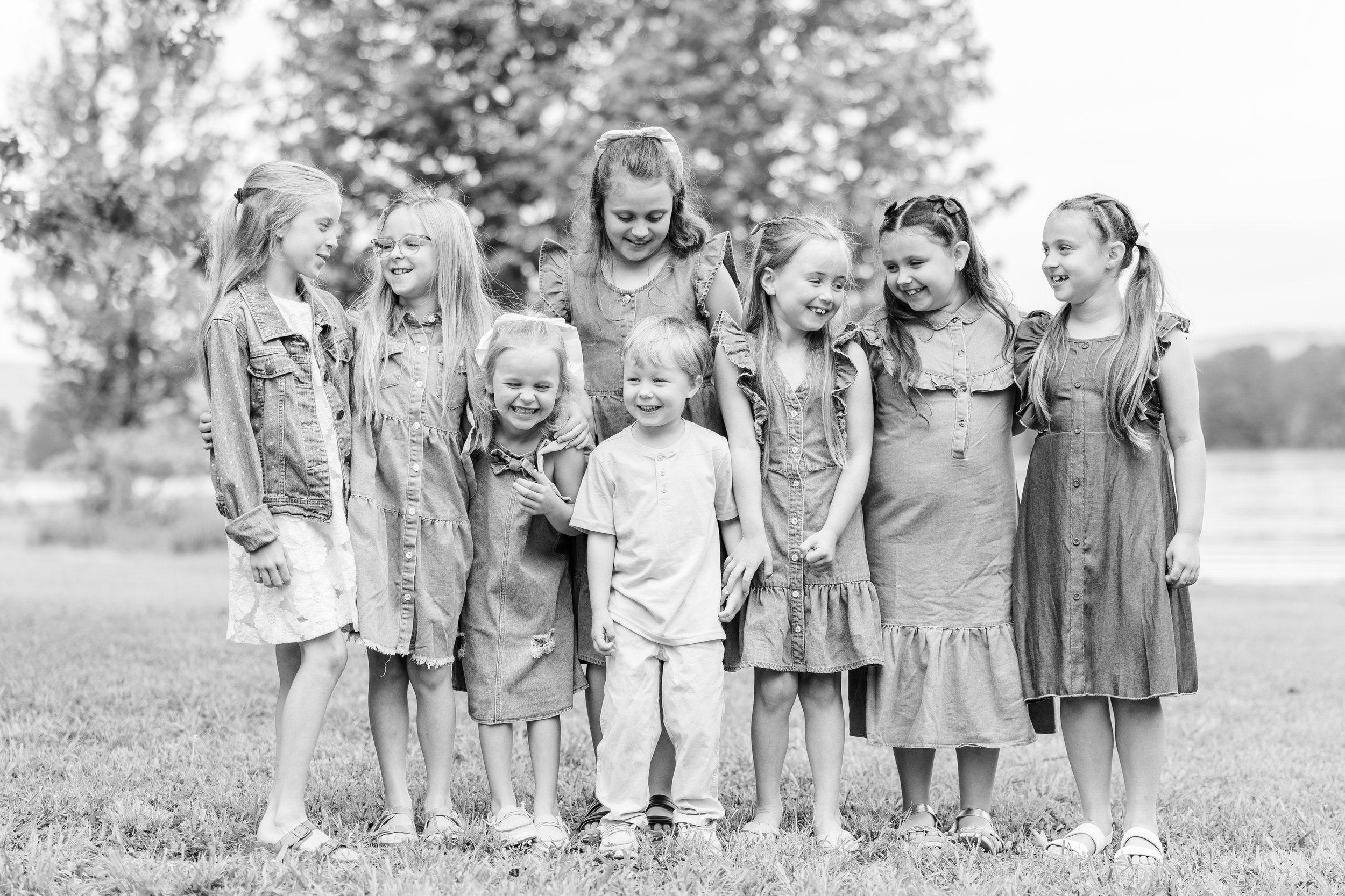 A group of nine young girls standing outdoors on grass, smiling and laughing together near a body of water with trees in the background.