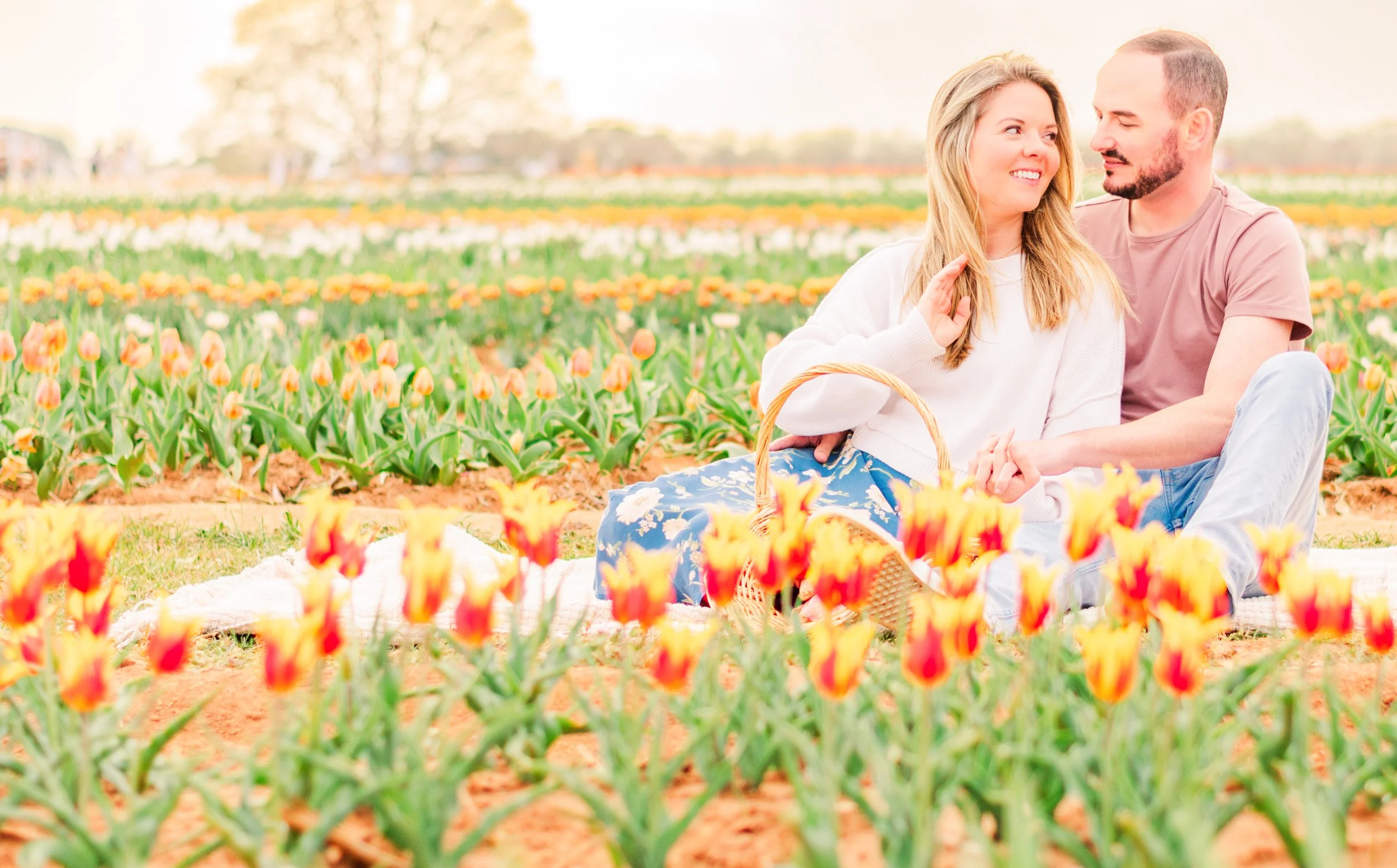 A young couple sitting on a blanket in a tulip field, enjoying each other's company, surrounded by orange and yellow tulips on a bright day.