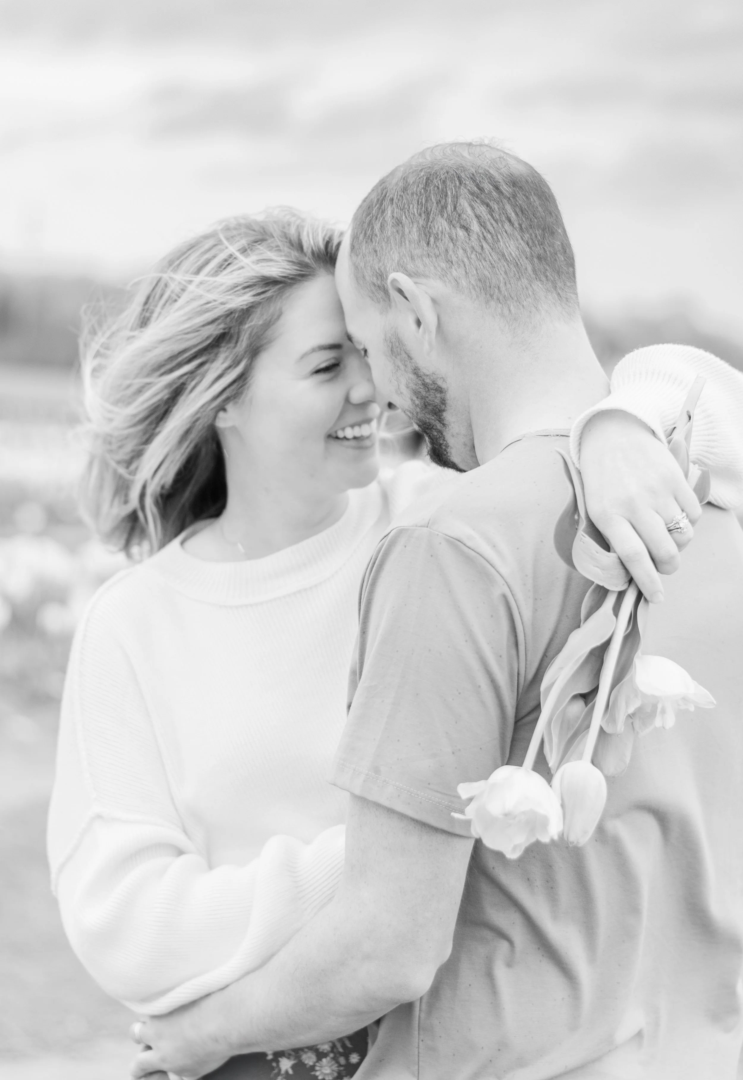 A black and white photo of a smiling woman and a man holding flowers close, with their foreheads touching, outdoors.