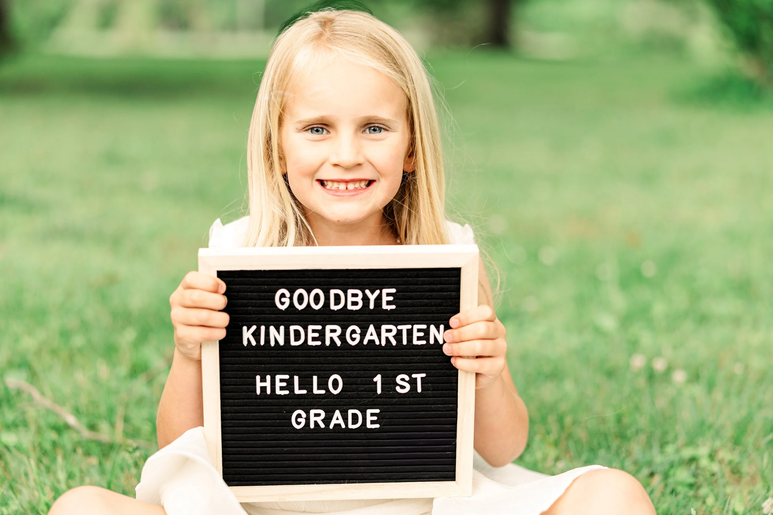 A young girl with blonde hair and a big smile sitting on grass outdoors, holding a black letter board with white letters that say "Goodbye Kindergarten Hello 1st Grade."