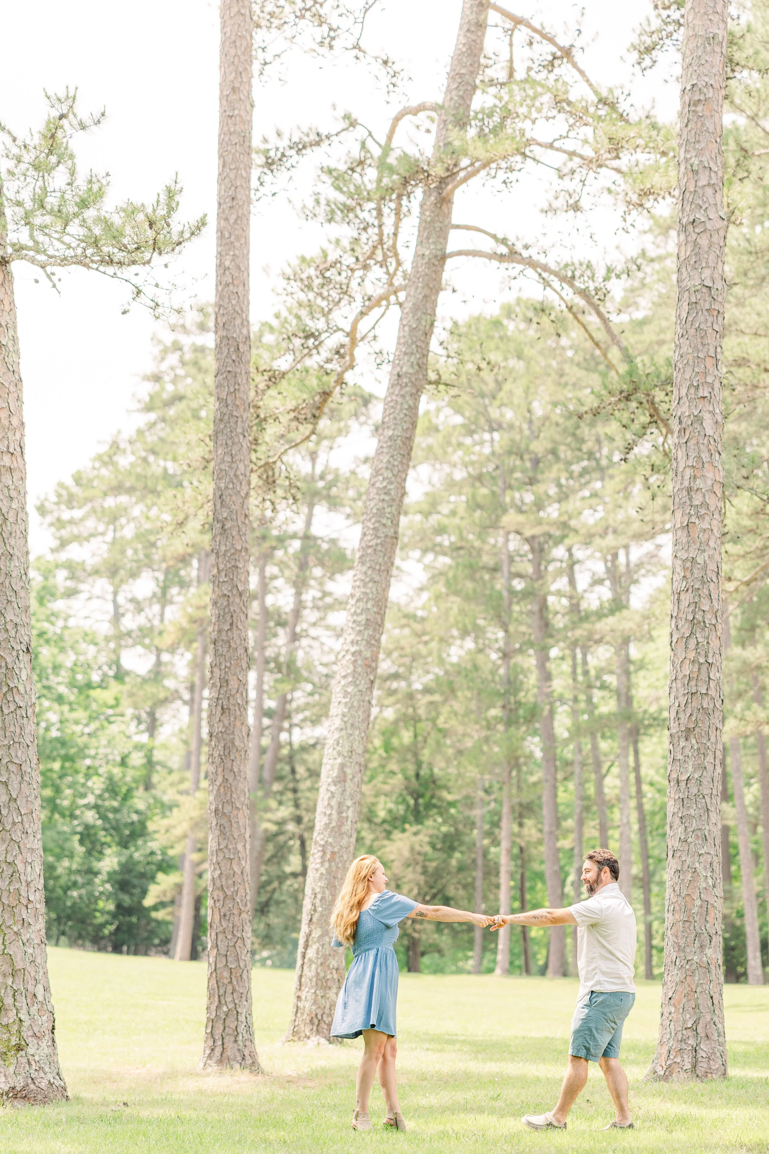 A couple holding hands and dancing in a forest clearing with tall trees and green grass.