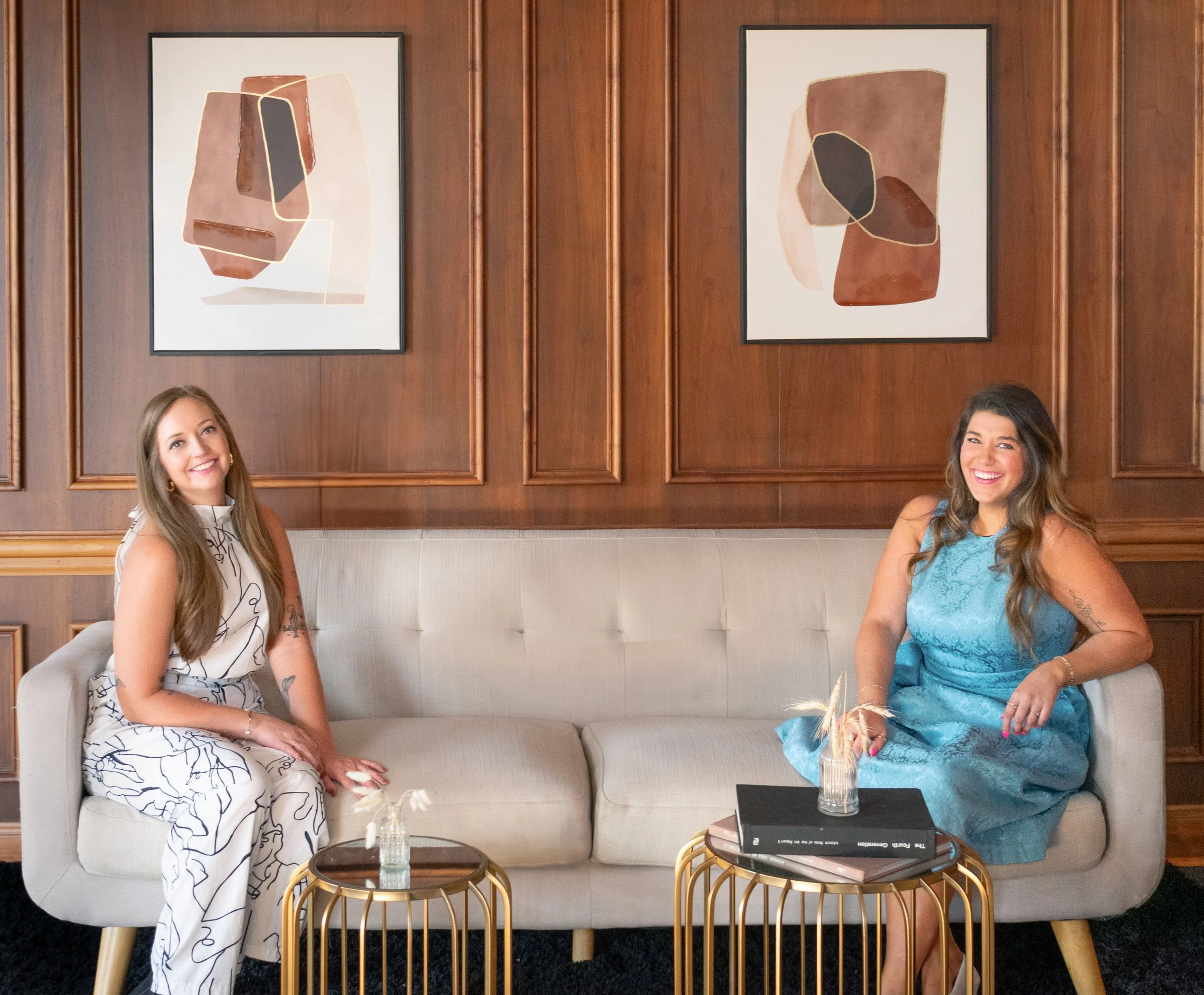 Two women sitting on a beige sofa with a wooden wall and abstract artwork behind them. One woman is wearing a white jumpsuit with black abstract lines, and the other is in a blue lace dress. They are smiling at the camera.