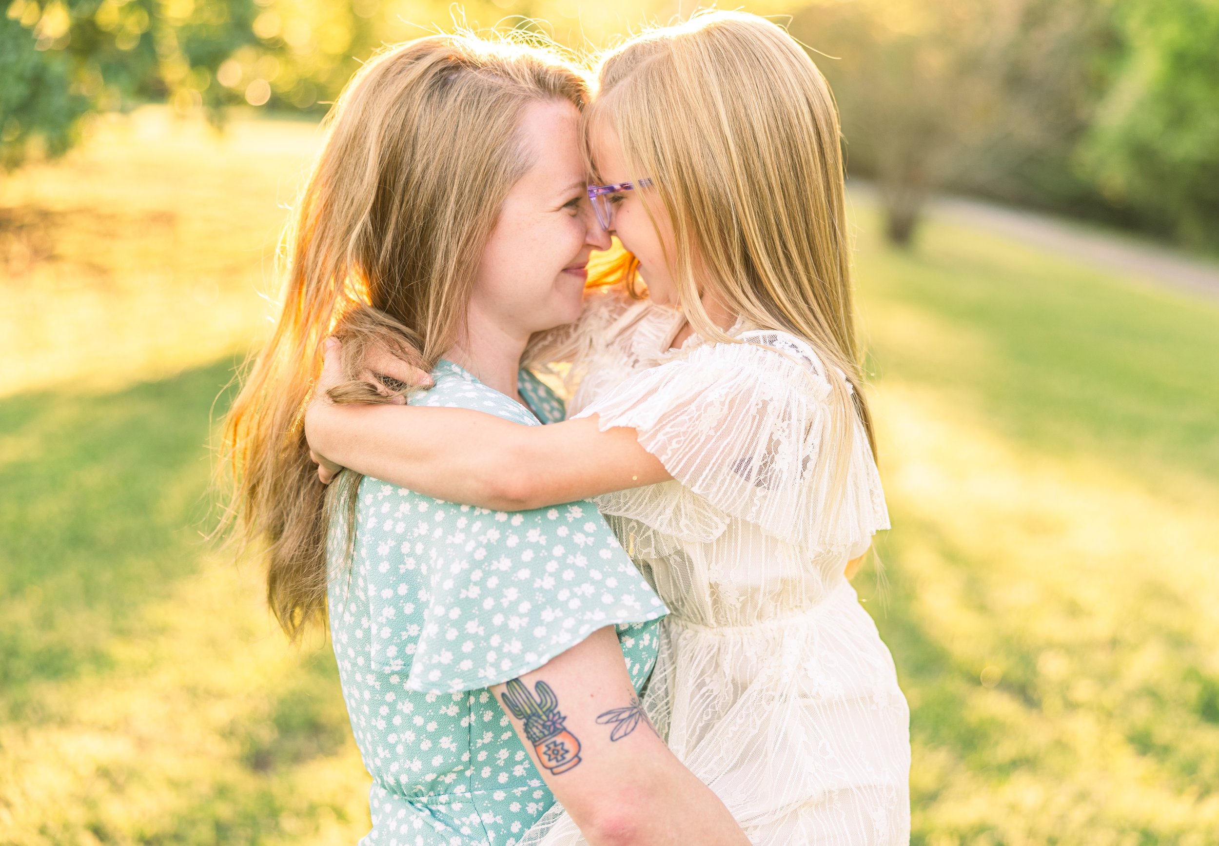 A woman and a young girl are outdoors, touching foreheads and smiling, with the woman holding the girl in her arms. The sunlight filters through trees in the background.