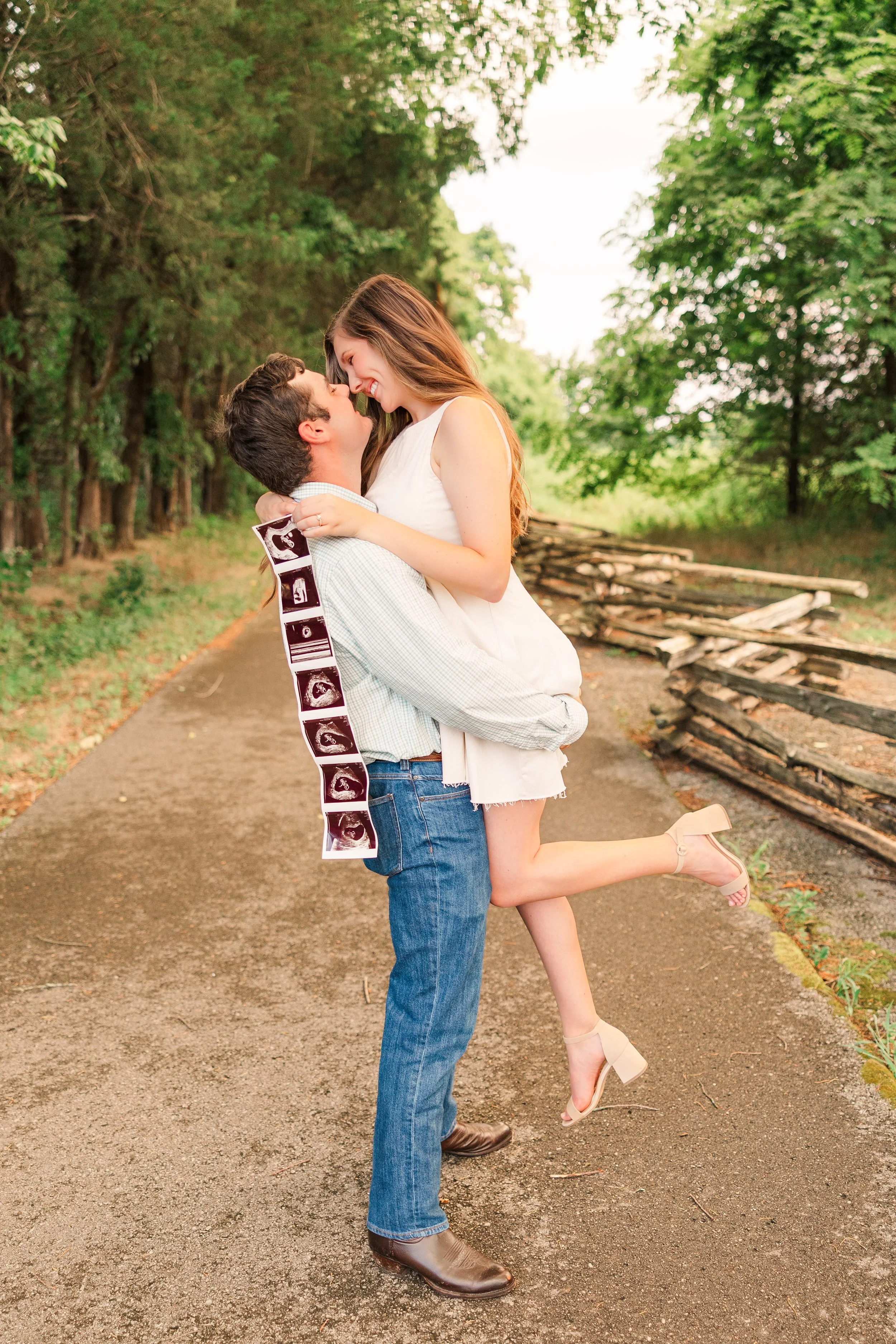 A couple standing on a dirt path outdoors, with the man lifting the woman who is holding ultrasound images, surrounded by green trees.