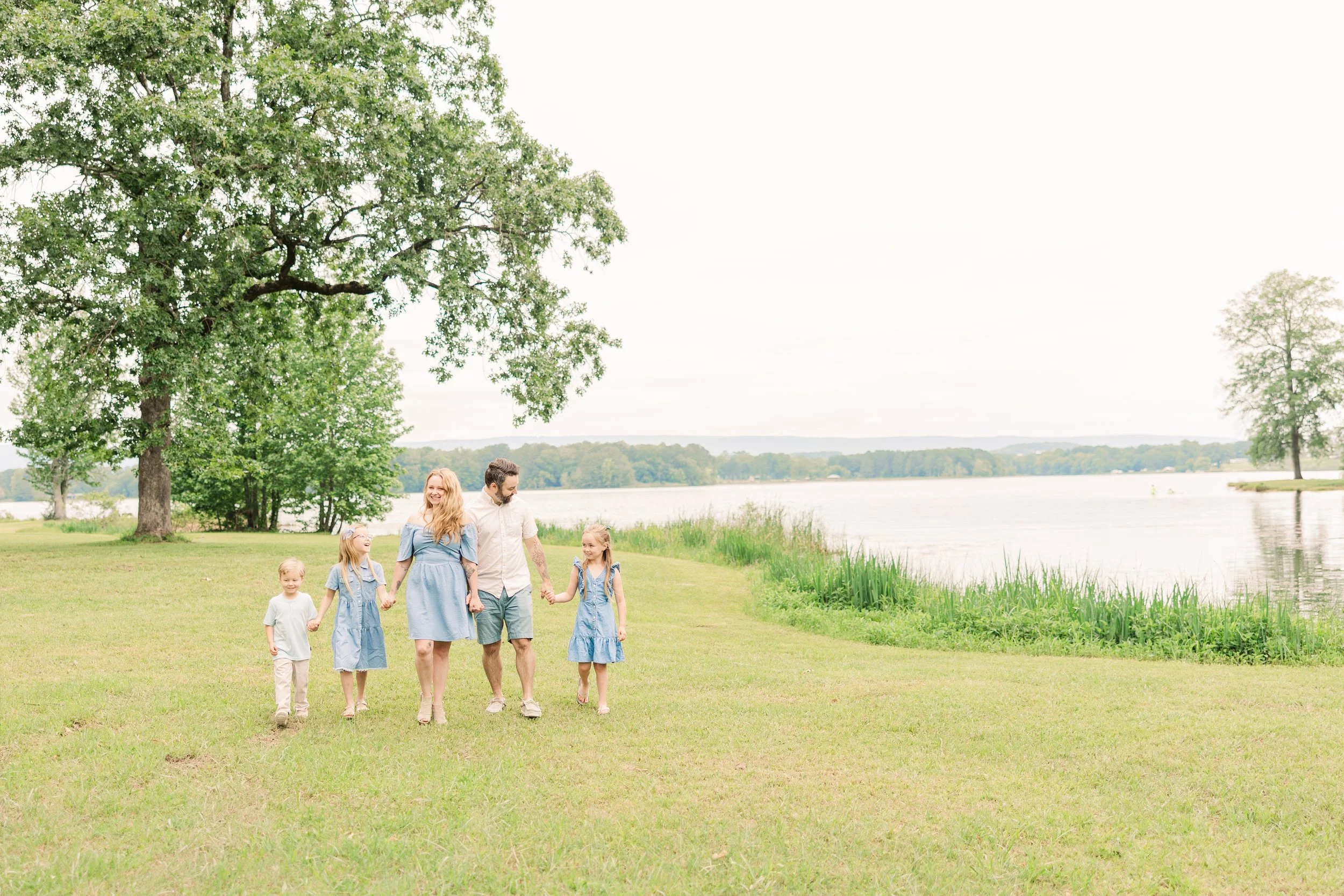 A family of six walking hand in hand near a lake, with trees and grassy shoreline in the background.