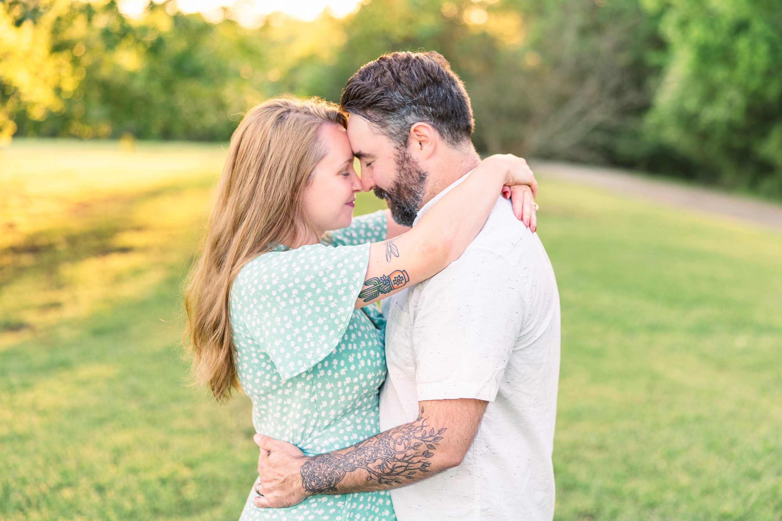 A couple embrace outdoors with their foreheads touching, eyes closed, in a grassy park with trees in the background during golden hour.